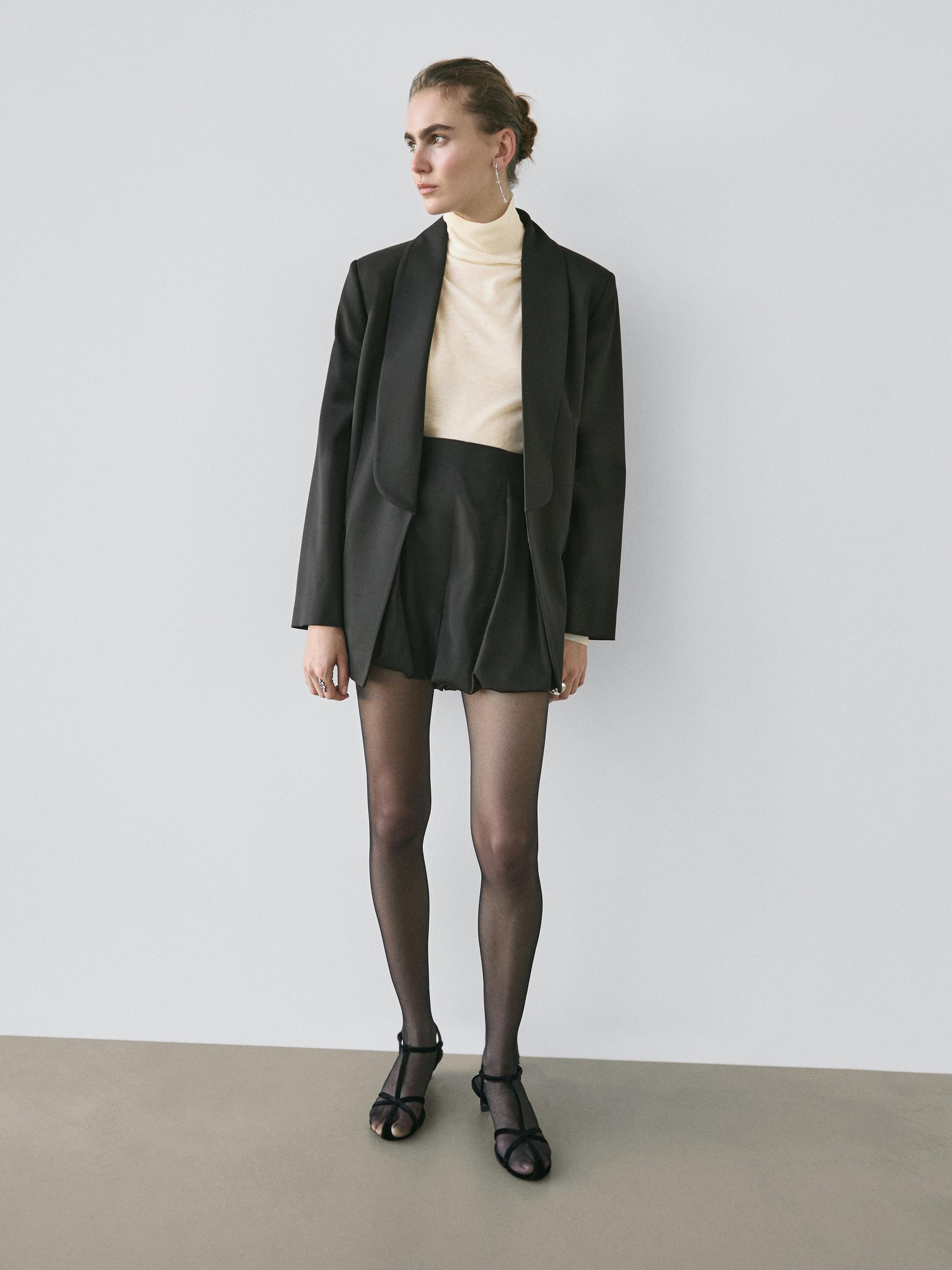 Ensemble blazer noir à revers, top beige à col roulé, short noir plissé et sandales noires.