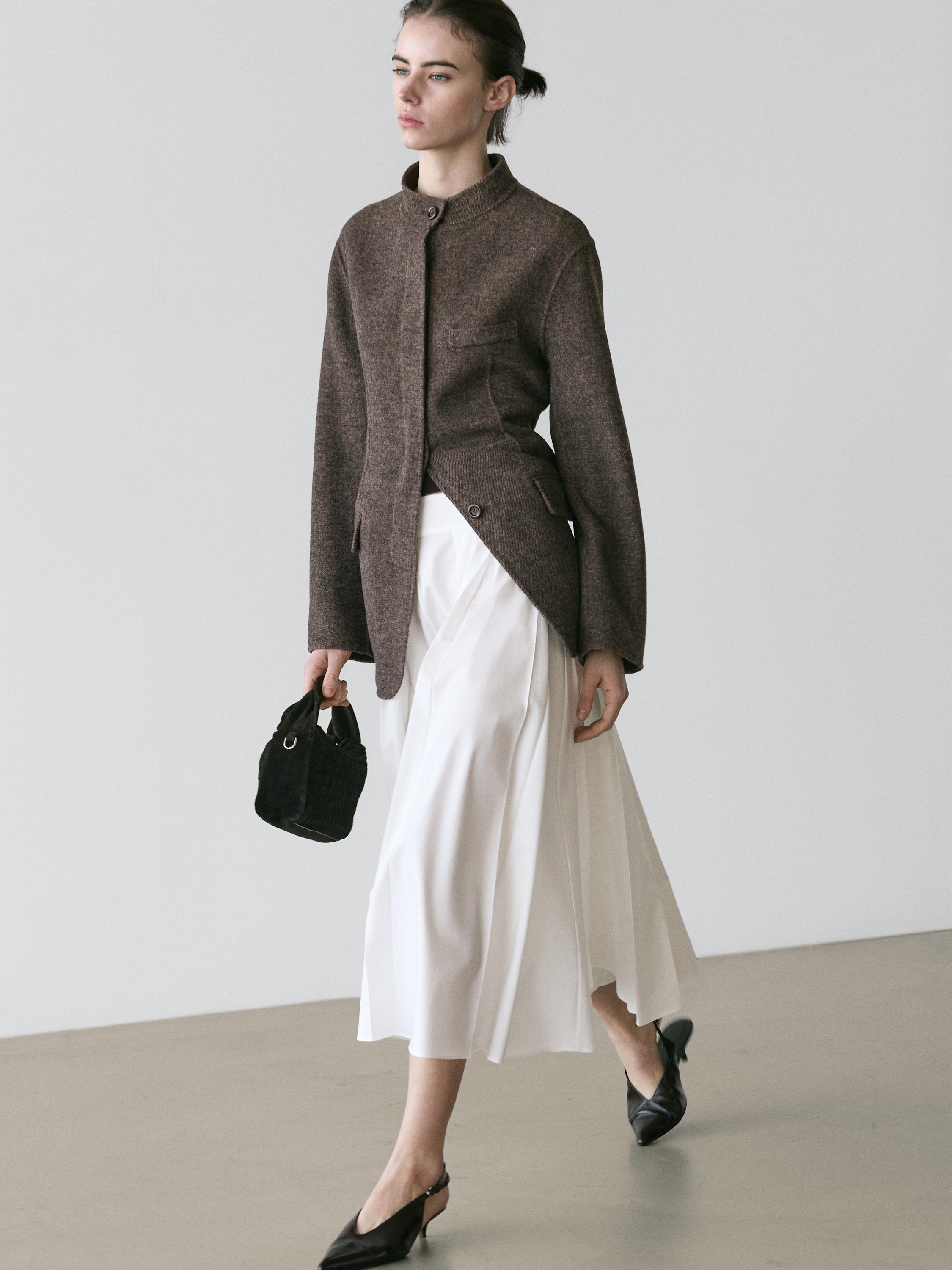 طقم جاكيت بني بجيوب وتنورة ميدي بيضاء بحلقات حزام، وحذاء أسود بكعب عالٍ وحقيبة يد سوداء.