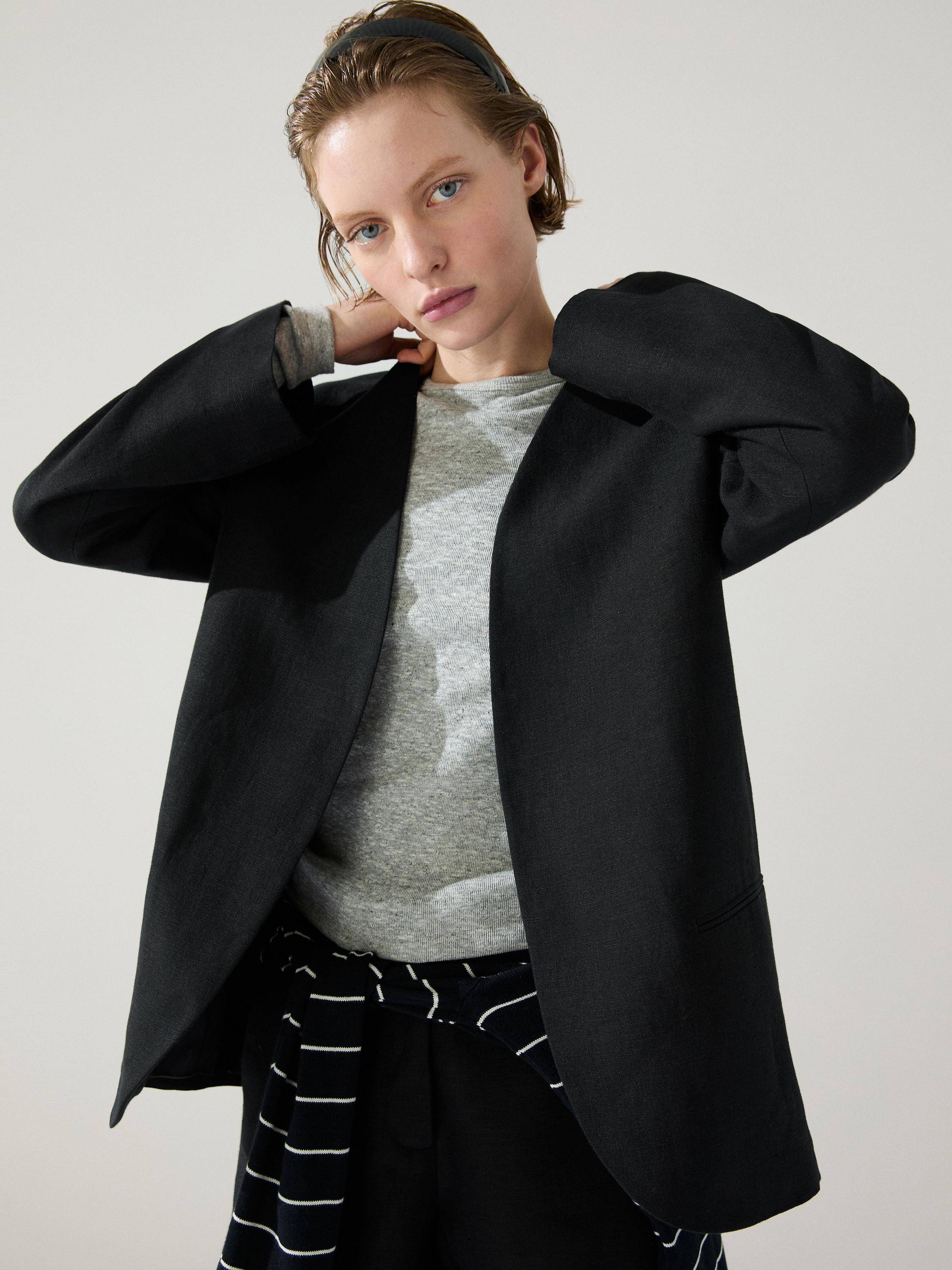 Półzbliżenie na czarną marynarkę bez klap, z bocznymi kieszeniami, na szarym T-shircie i czarnych spodniach z białymi paskami.