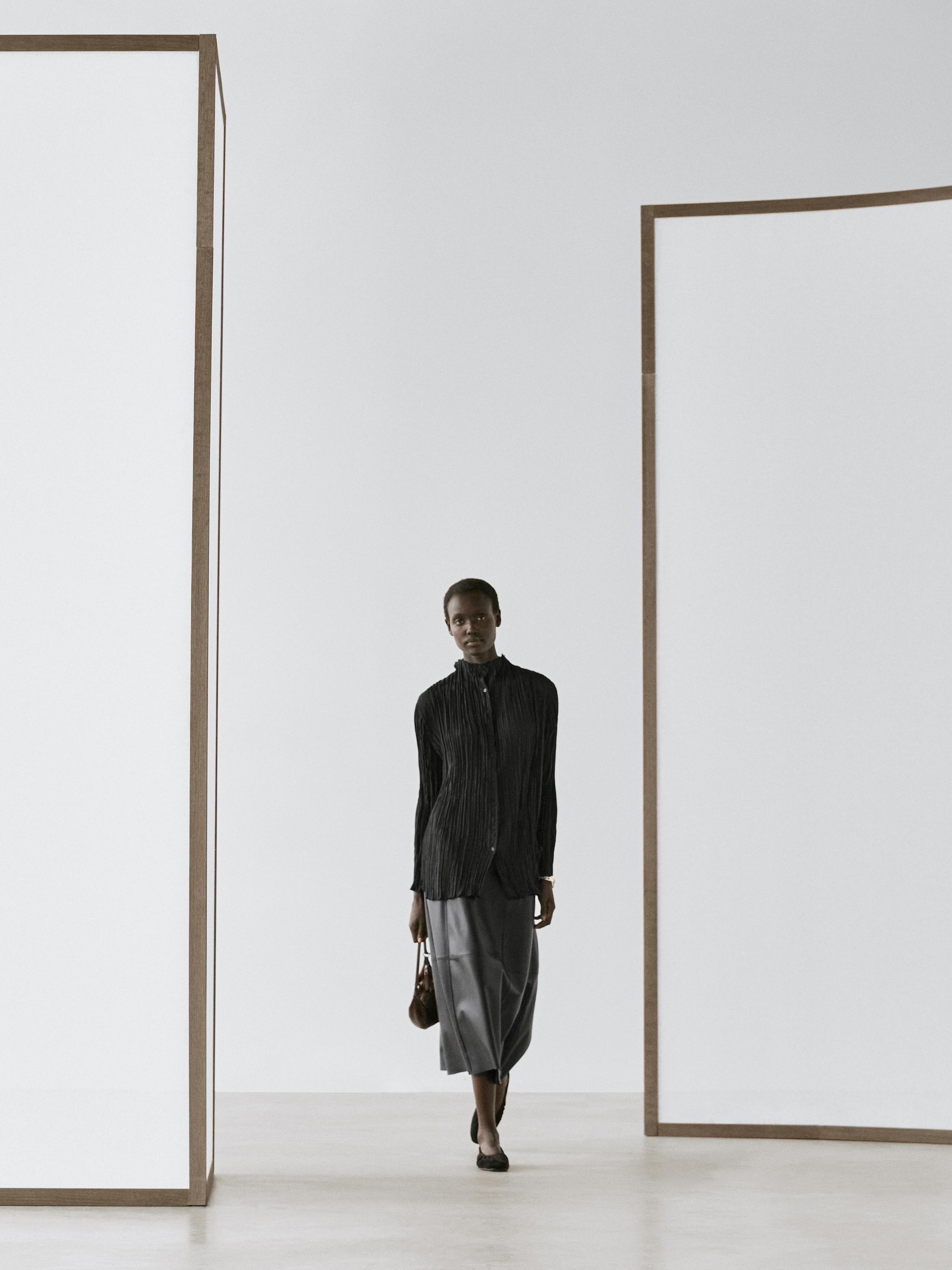 Ensemble chemise noire plissée et jupe midi grise, avec petit sac et chaussures plates noires.