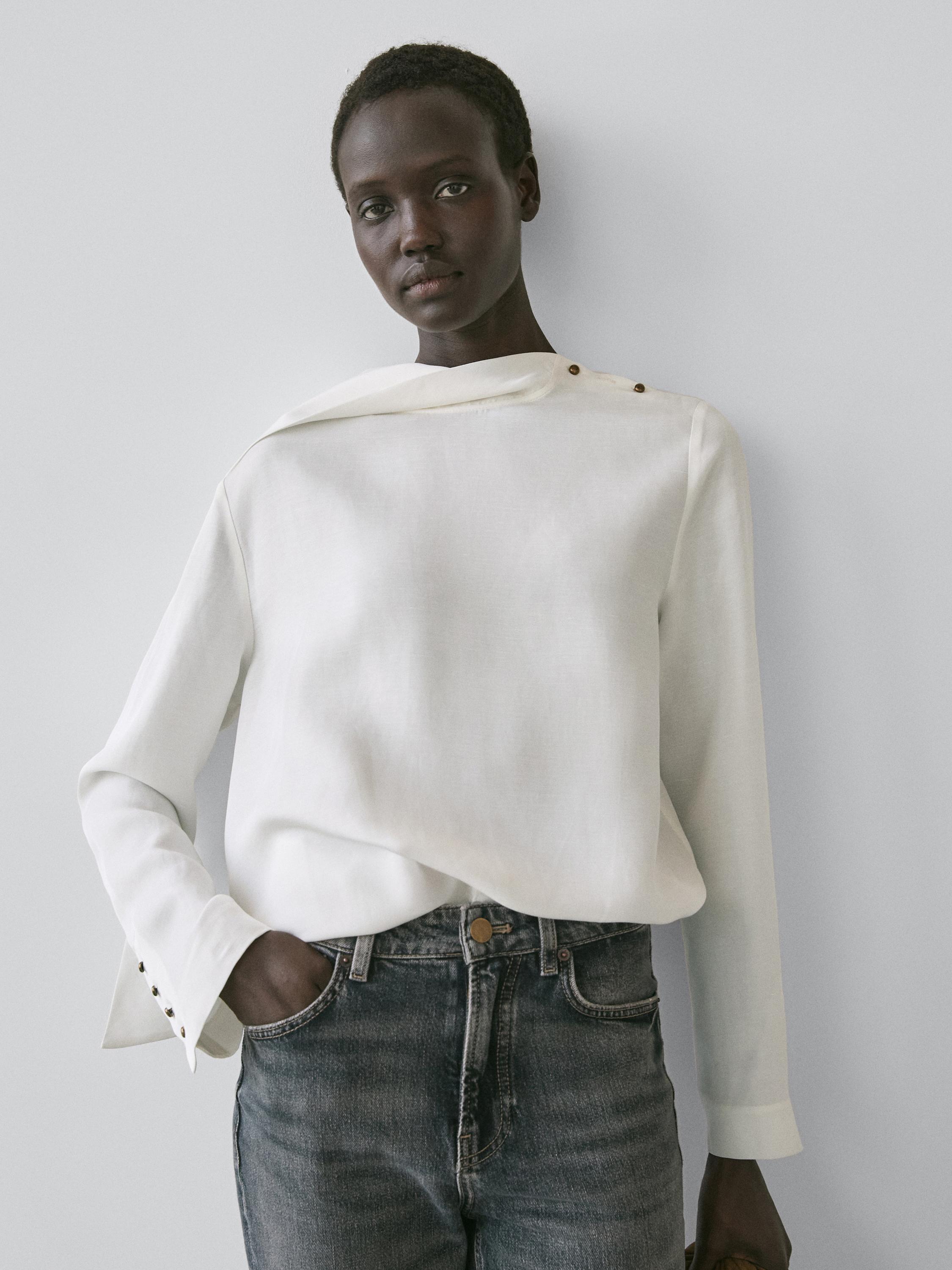 Pamje nga afër e këmishës së bardhë me kopsa në shpatull dhe mansheta, e kombinuar me pantallona xhins të errët.