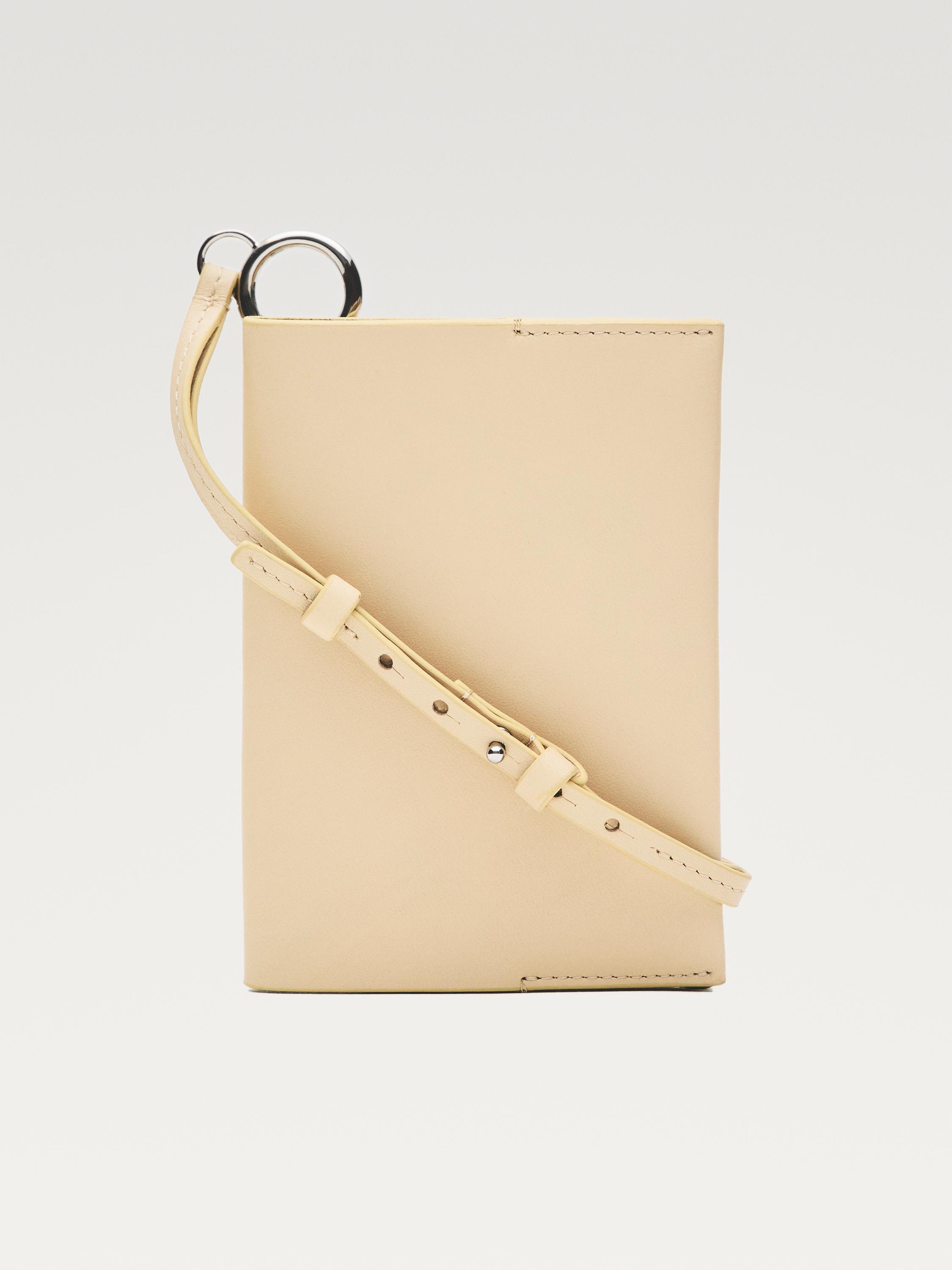 Vista frontal de porta-moedas baunilha com alça ajustável e argola metálica na parte superior.