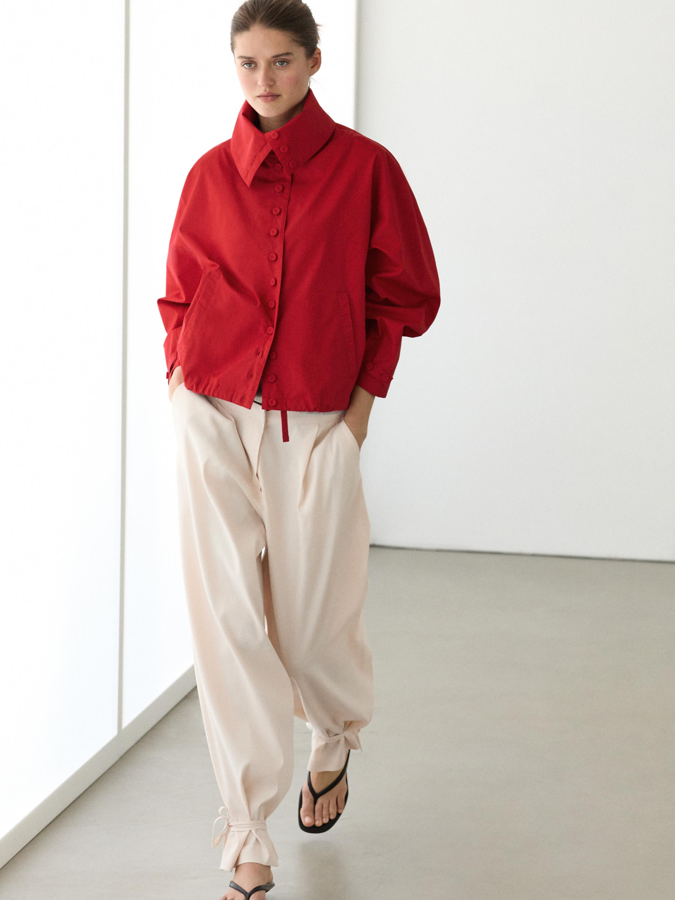 Rood jasje met knopen en col, losse beige broek met strikjes bij de enkels en zwarte sandalen.