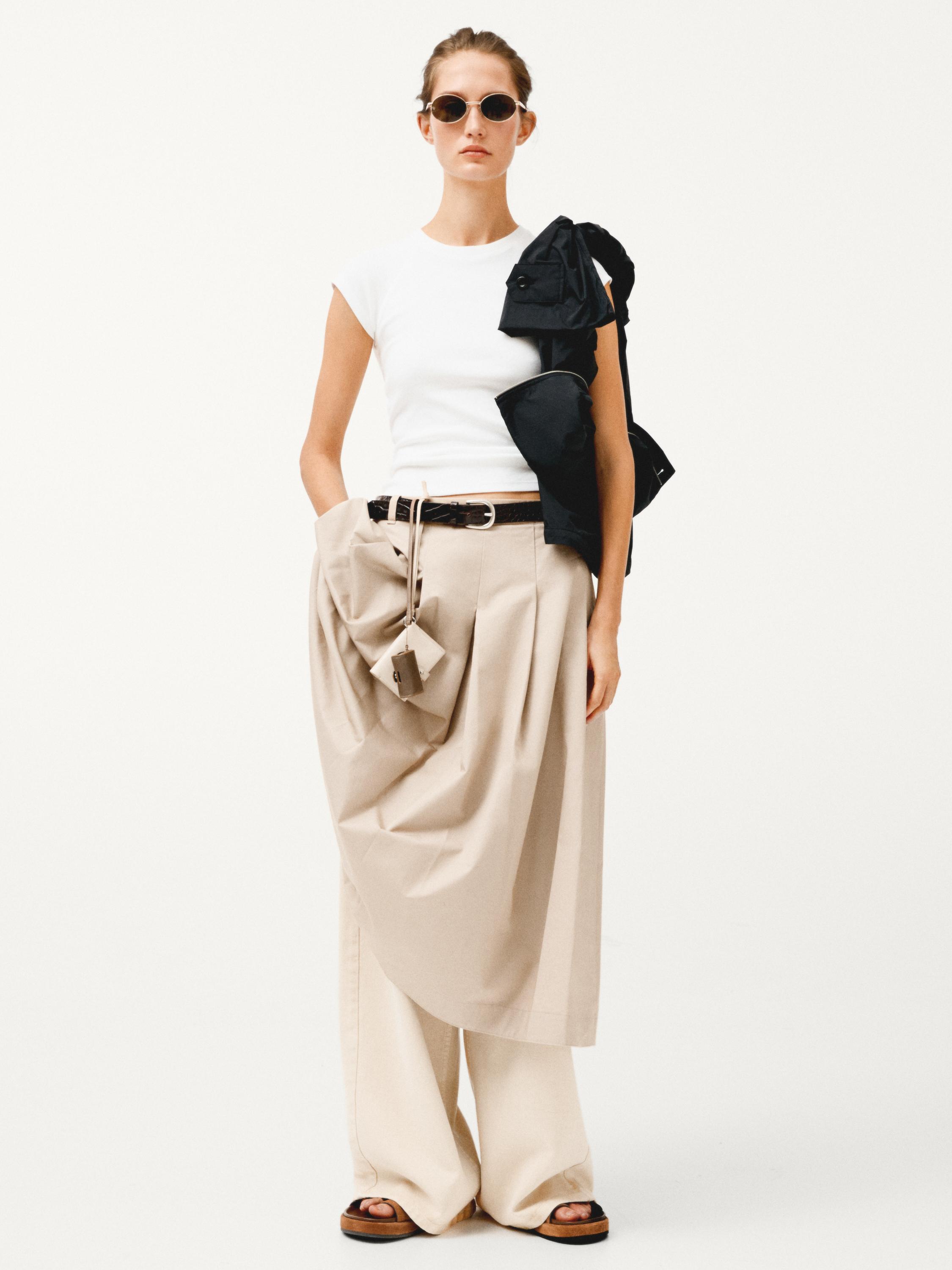 Ensemble jupe midi plissée couleur sable, pantalon large et débardeur blanc. Ceinture noire visible.