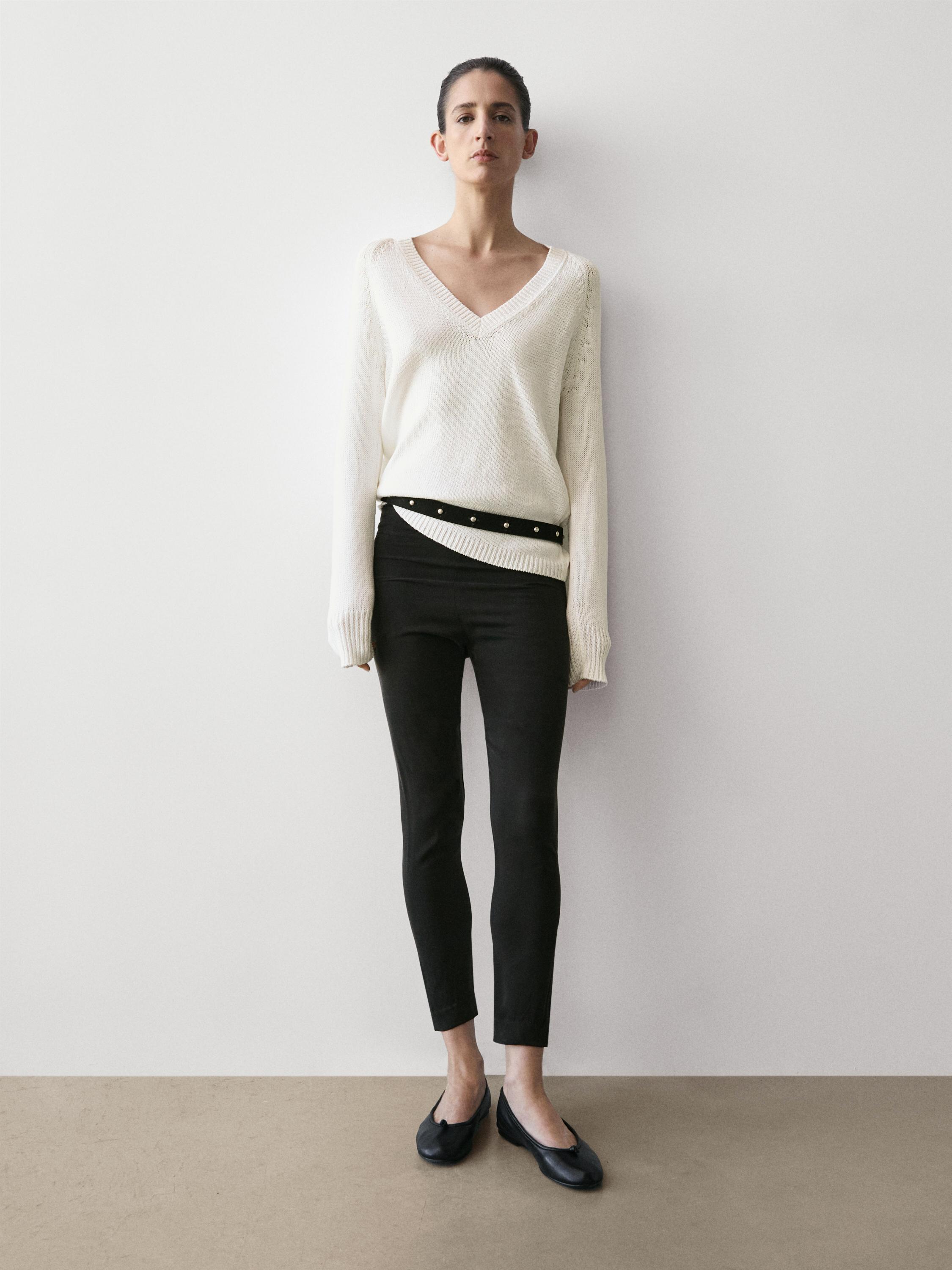 Conjunt de suèter blanc de coll en V i pantalons legging negre ajustat, amb cinturó decoratiu i sabates planes negres.