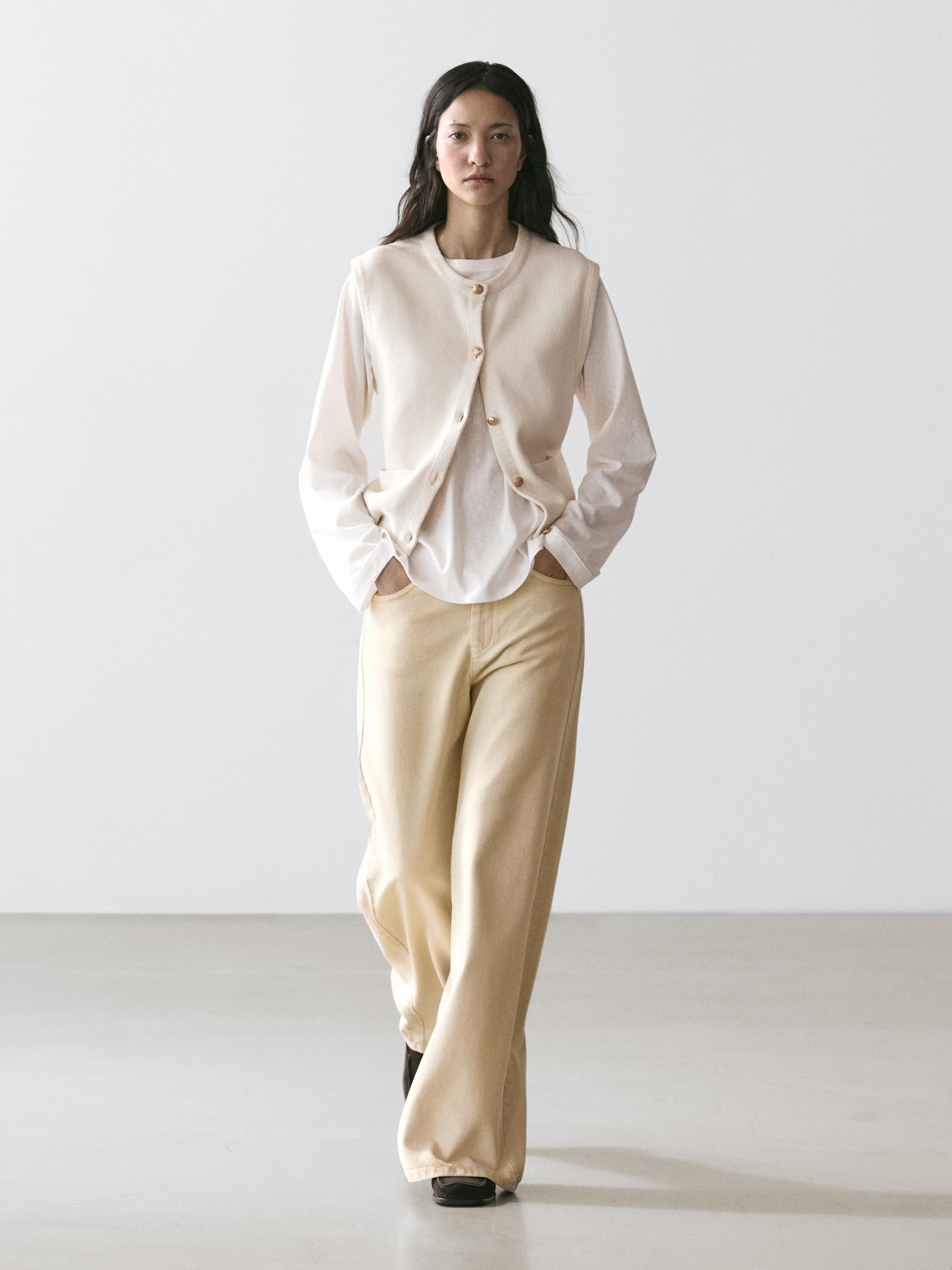 Gordina koloreko txaleko botoi eta poltsikoduna, kamiseta zuri bat eta galtza beix zabalak.