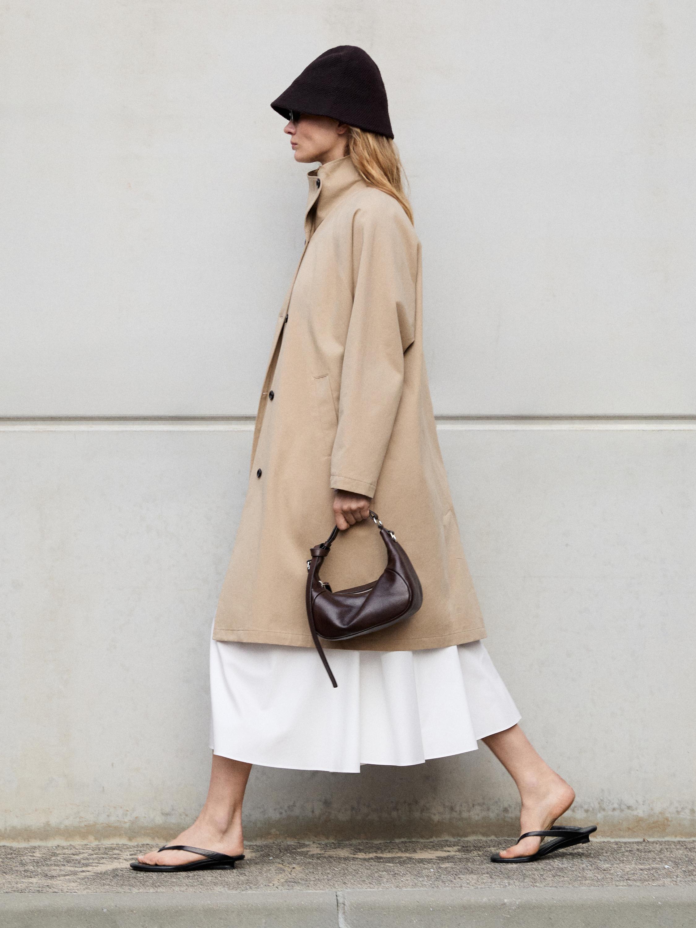 Yandan bağlamalı uzun bej trençkot takımı, midi beyaz etek, kahverengi çanta ve siyah sandaletler.