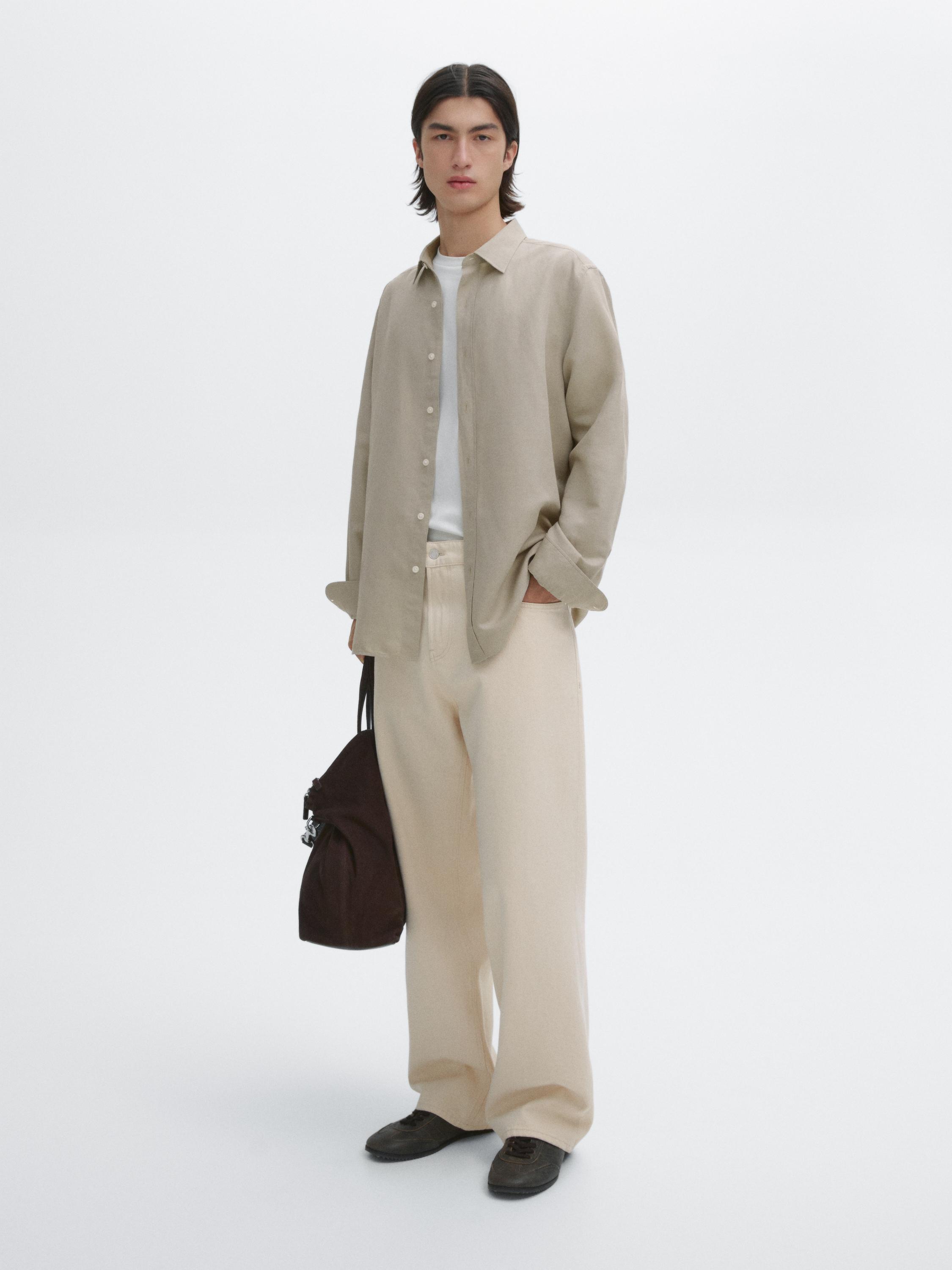 طقم مكون من قميص بيج، وقميص أبيض، وبنطلون بيج واسع وحذاء داكن، مع حقيبة بنية اللون.