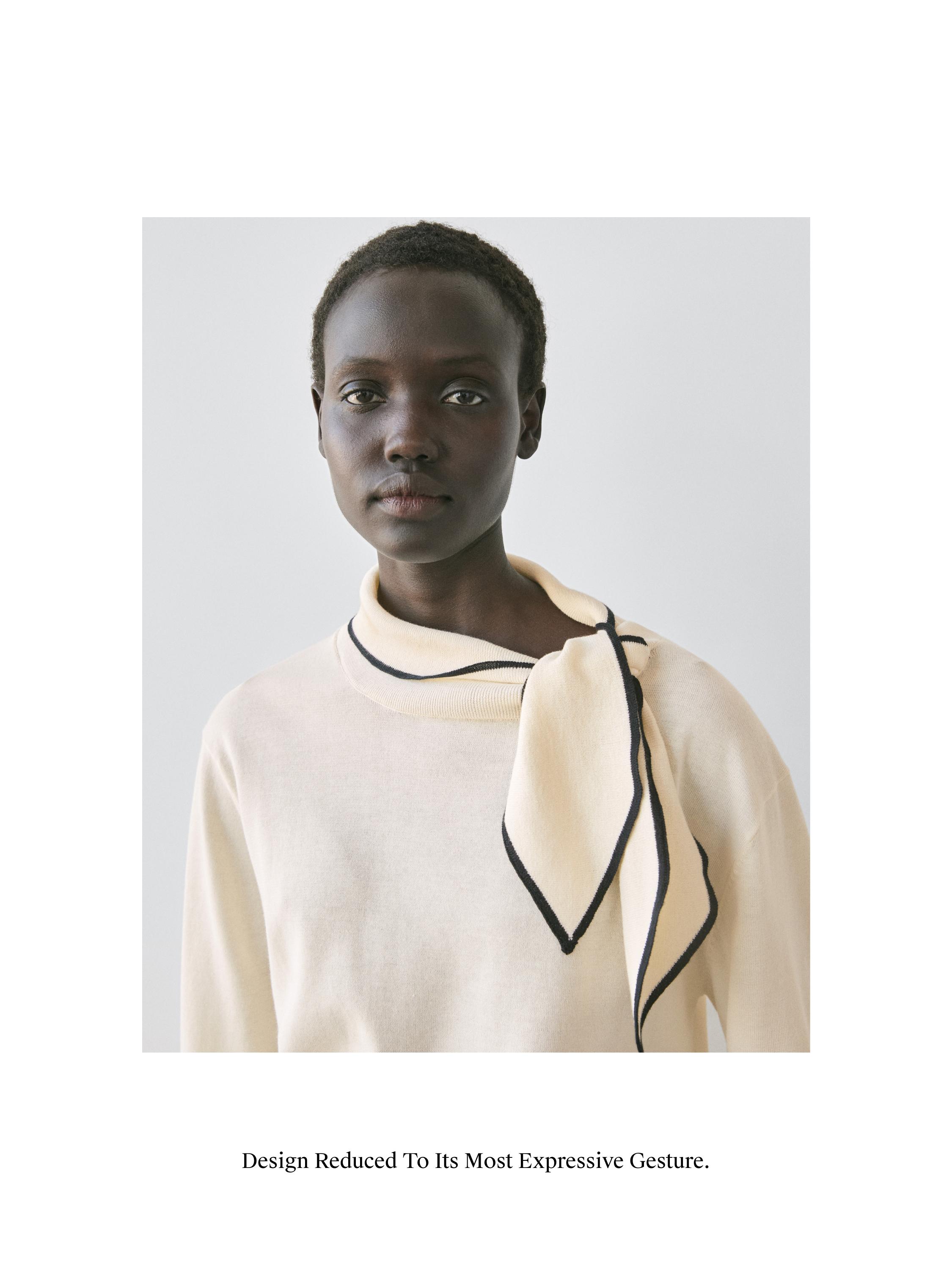 plaintext 스카프 디테일이 있는 아이보리색 스웨터의 중간 클로즈업 샷. 이미지 텍스트: "Design Reduced To Its Most Expressive Gesture."