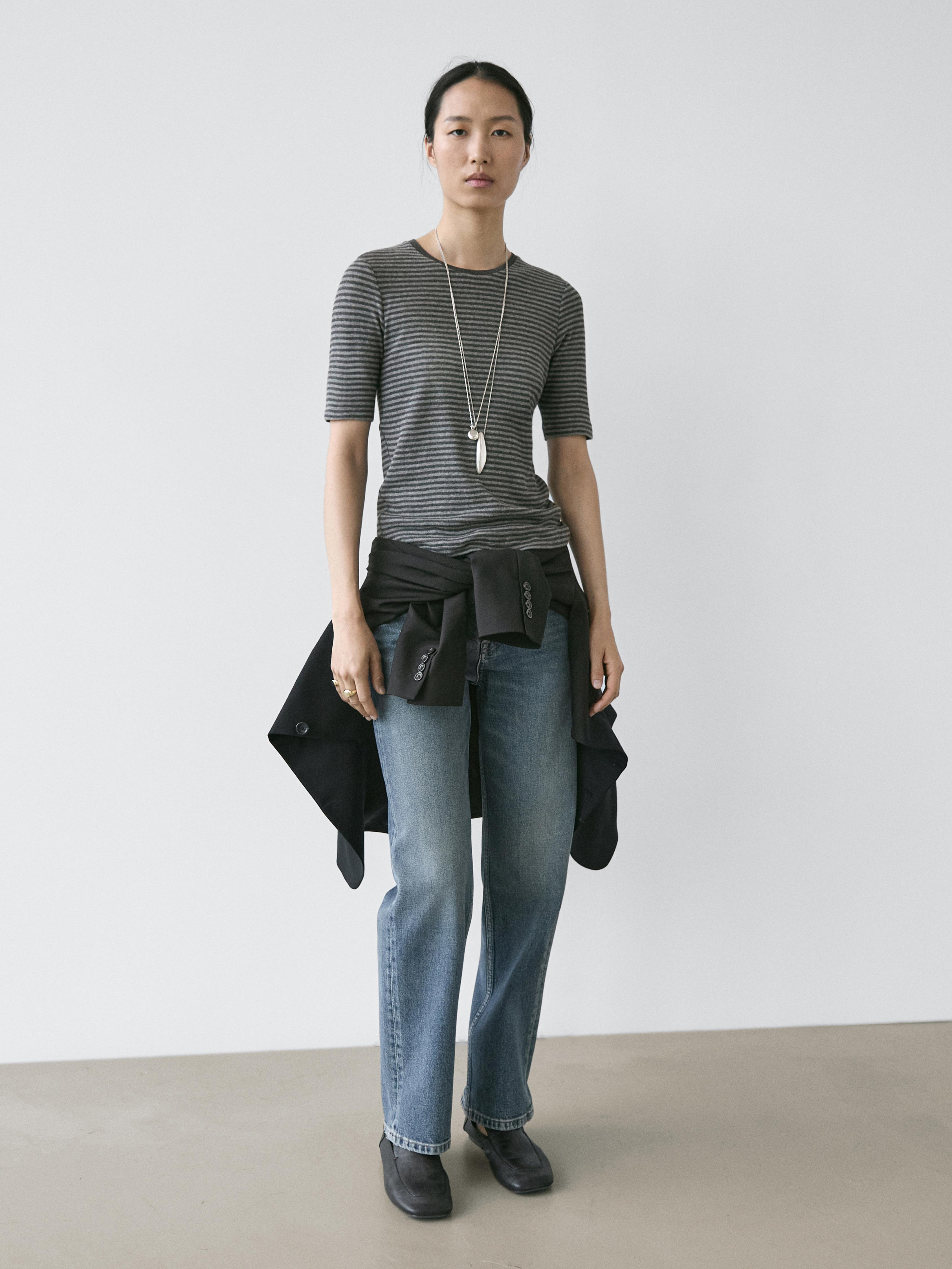 Conjunto de camiseta gris de rayas manga corta, jeans azules rectos y botas negras. Chaqueta negra atada a la cintura.