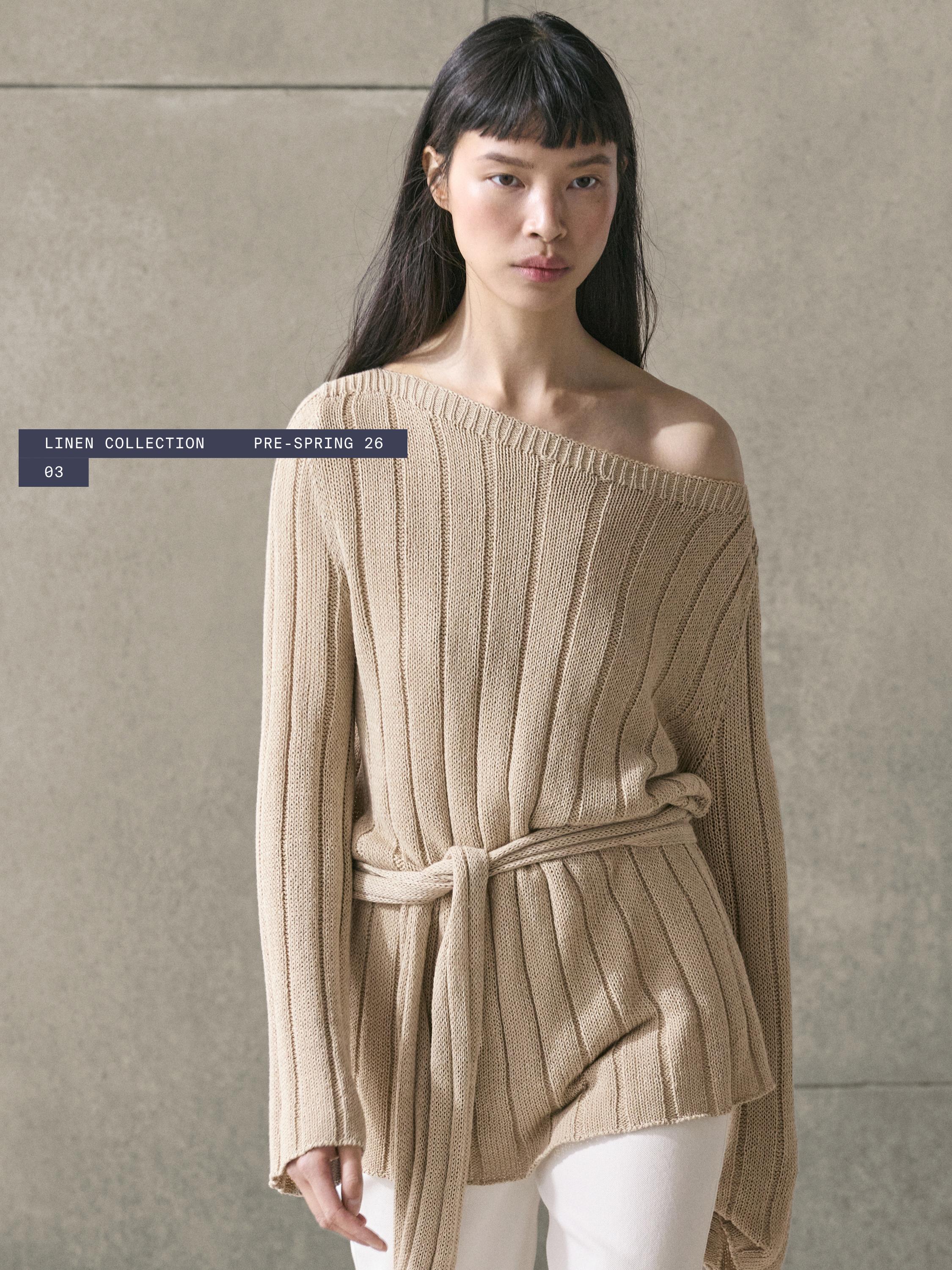 Комплет беж плетен џемпер со бродско деколте и ремен, текст "LINEN COLLECTION PRE-SPRING 26 03", бели панталони.