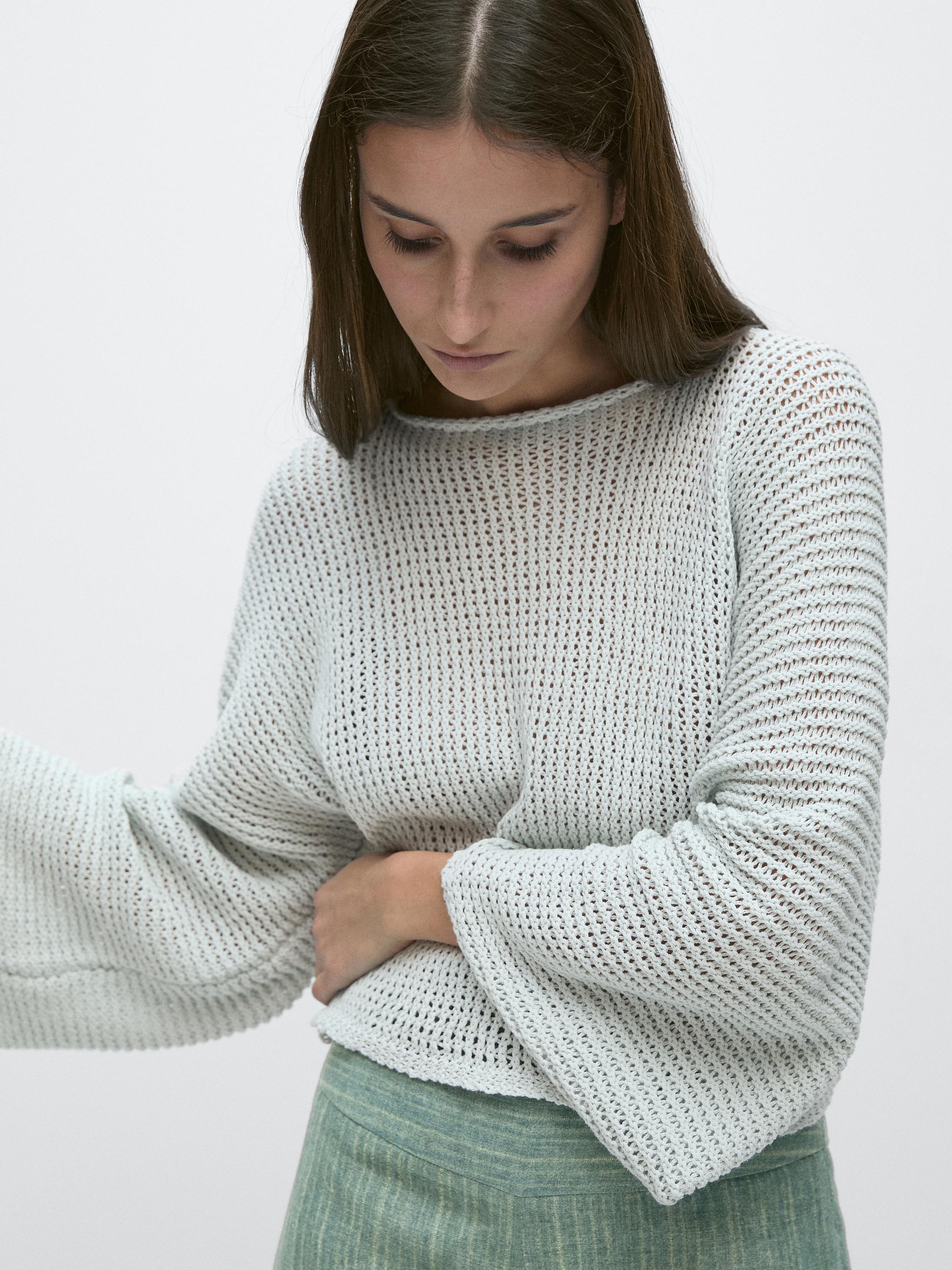 Plano medio de suéter blanco de punto calado con mangas largas y cuello redondo. Falda verde visible en la parte inferior.