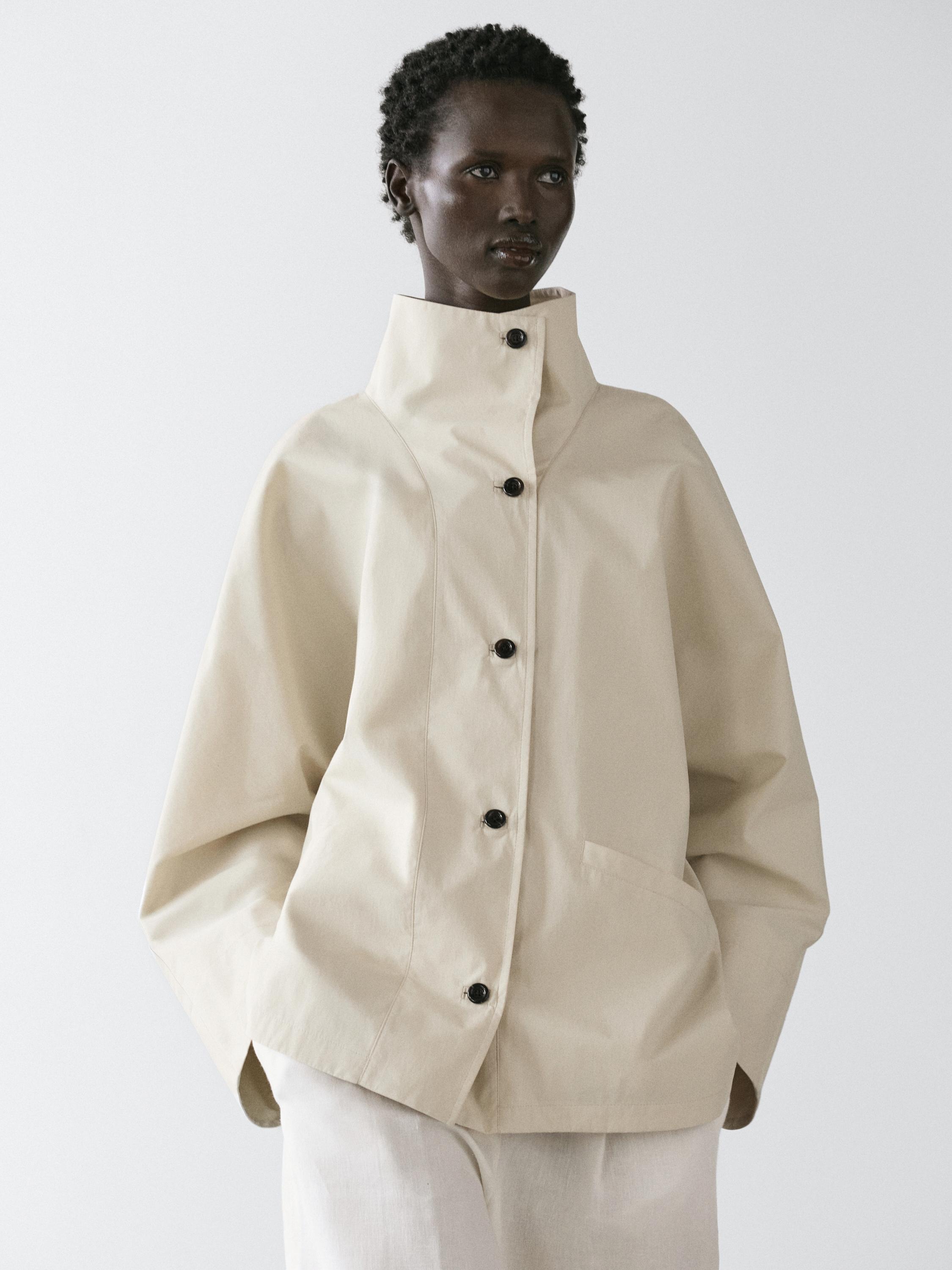 Rauwe jackset met opstaande kraag en zichtbare zwarte knopen, zijzakken, over een witte broek.