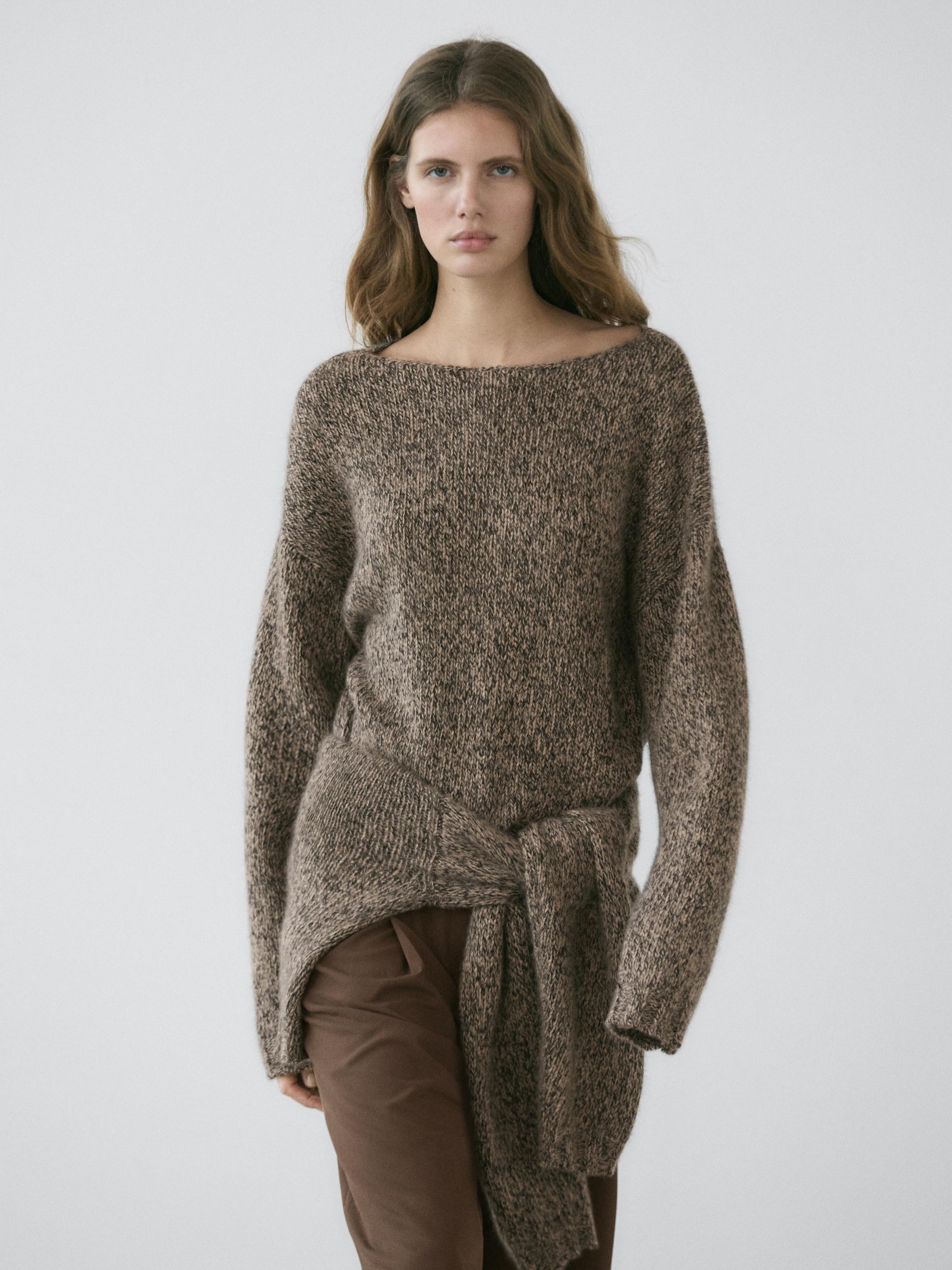 Sett med tykkstrikket minkfarget genser med knyting i midjen og brune bukser.