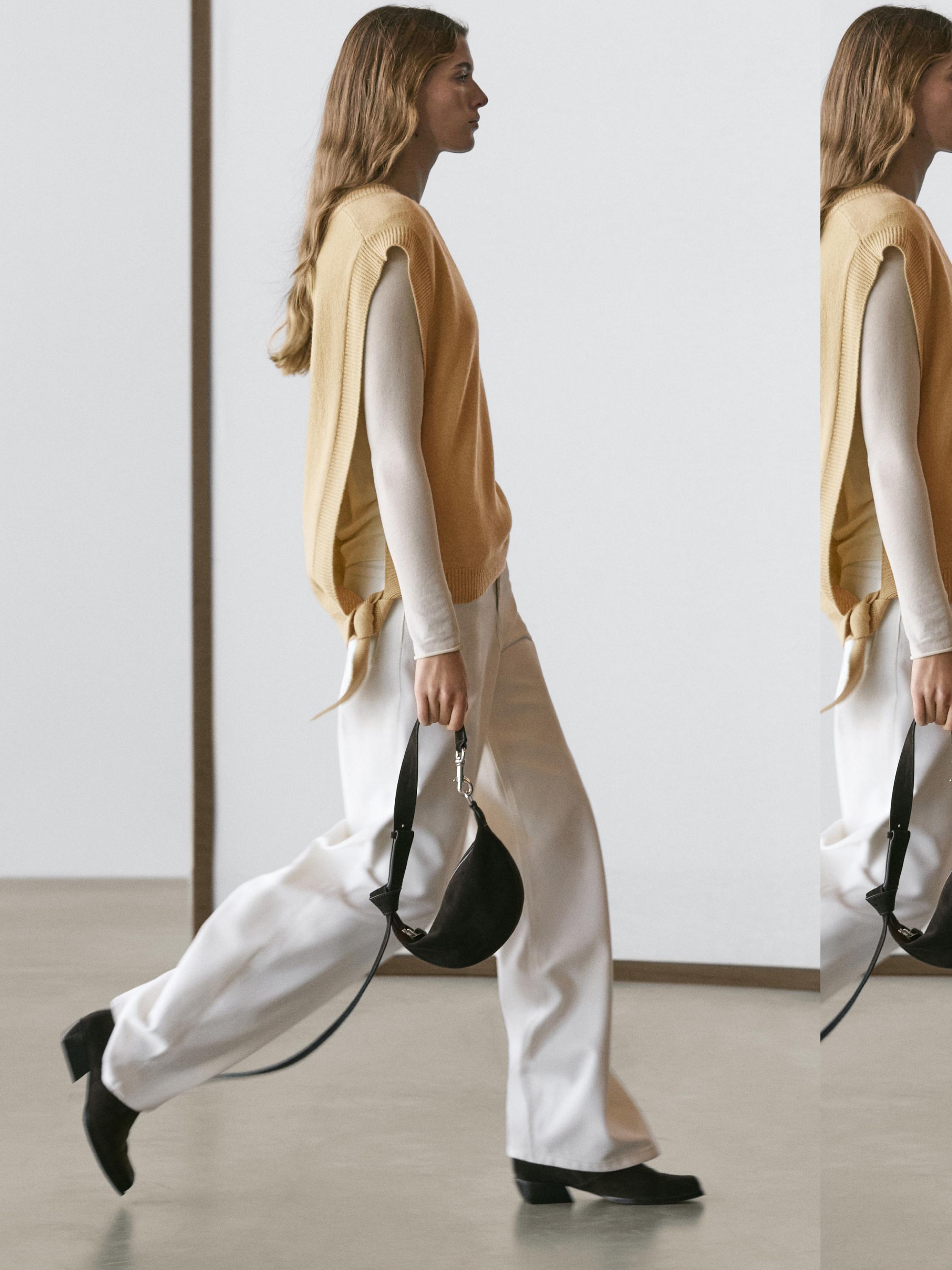 Varrukateta kollane vest, pikkade varrukatega beež pluus, laiad valged püksid ja must kott.