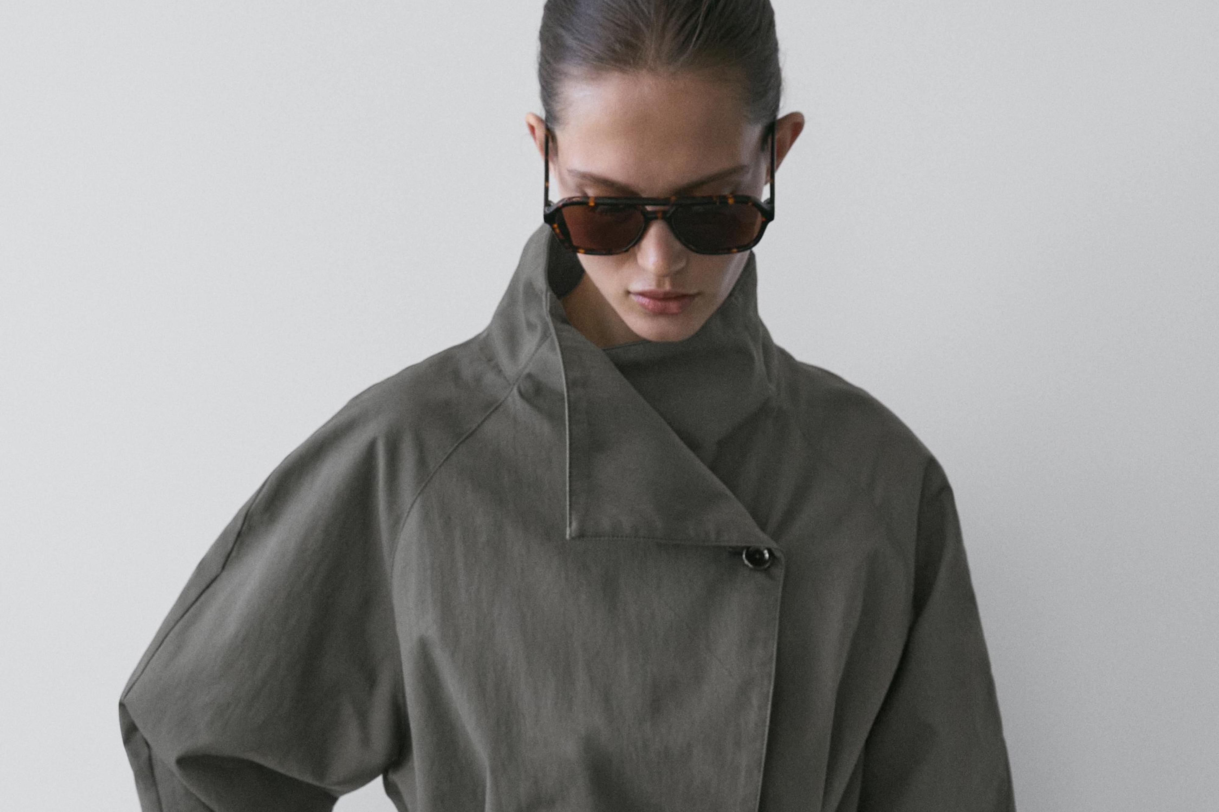 لقطة متوسطة لمعطف ترنش قصير أزرق متوسط مع حزام وياقة عالية غير متماثلة، زر ظاهر في الجزء العلوي.