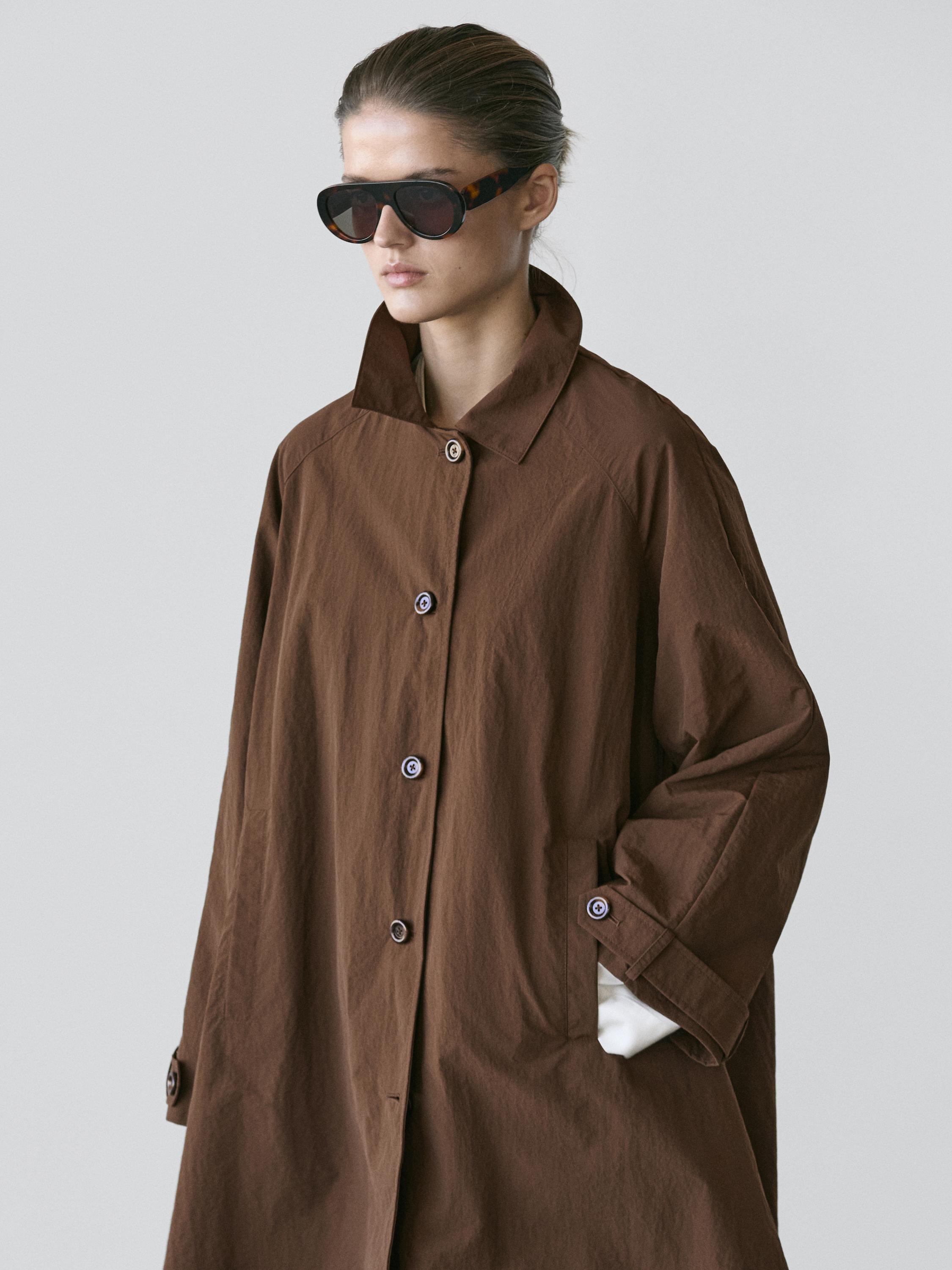 plaintext Srednji posnetek rjavega plašča z visokim ovratnikom, sprednjim zapenjanjem na gumbe in stranskimi žepi.