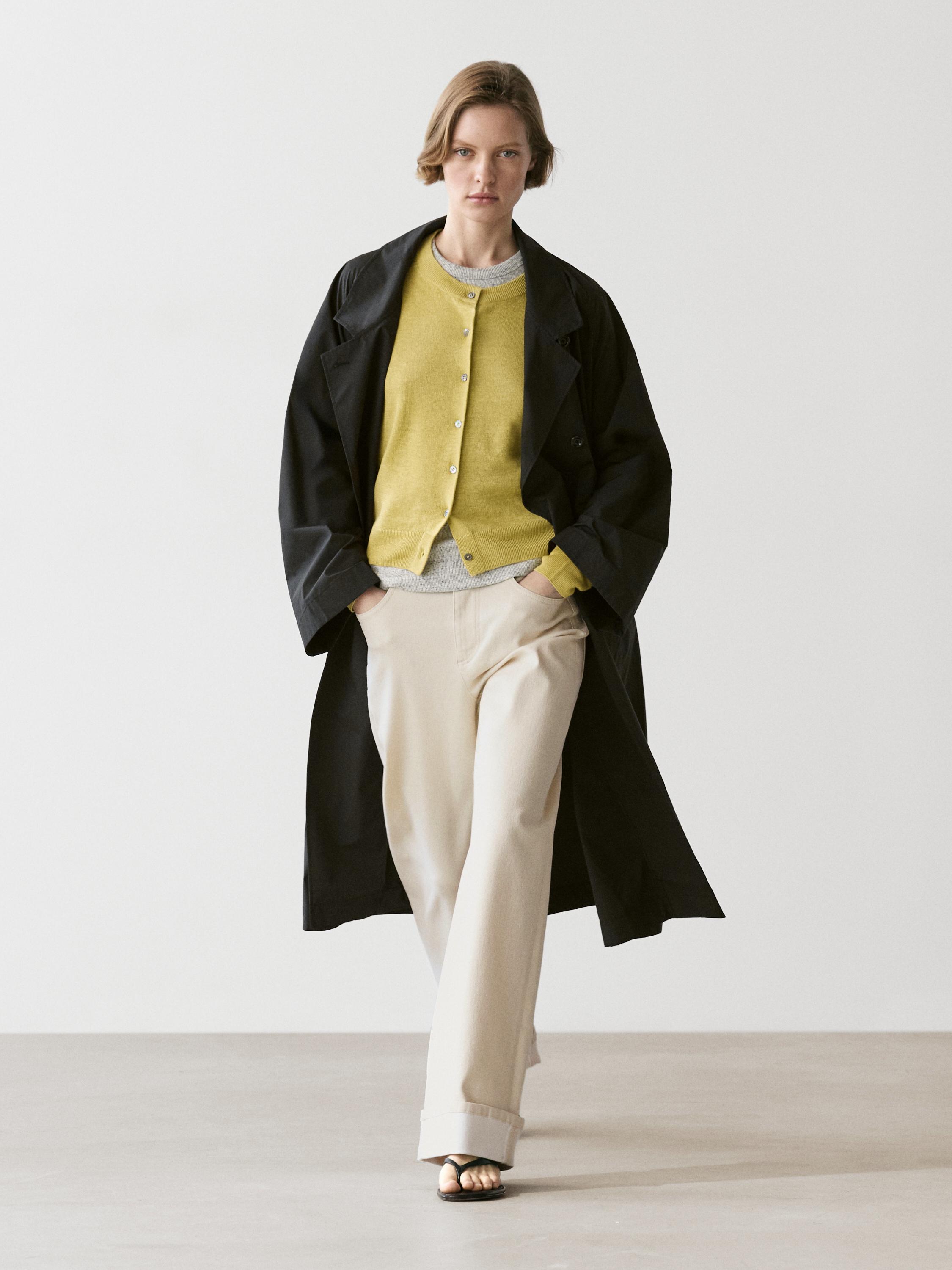 Limegrünes Cardigan-Set mit Knöpfen, langer schwarzer Mantel, beigefarbene Hose und schwarze Sandalen.