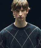 عارضة أزياء مرتدية سويتر بنقشة ماسية داكنة على خلفية سوداء. شعرها بني ولها تعبير جدي.