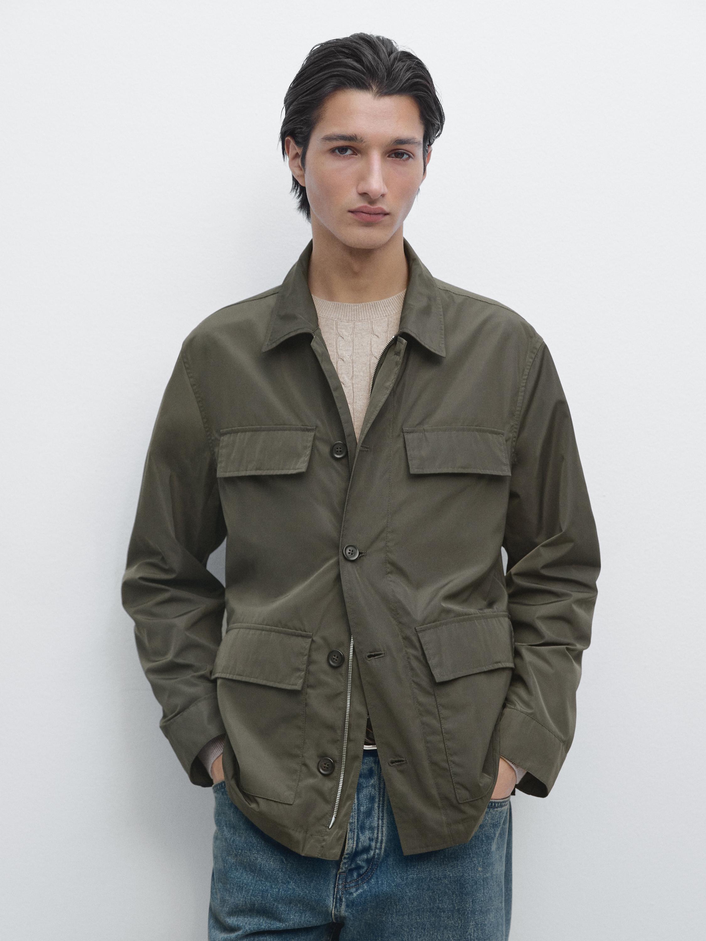 Ensemble composé d'une veste saharienne kaki avec poches frontales et boutons, sur un pull beige et un jean bleu.