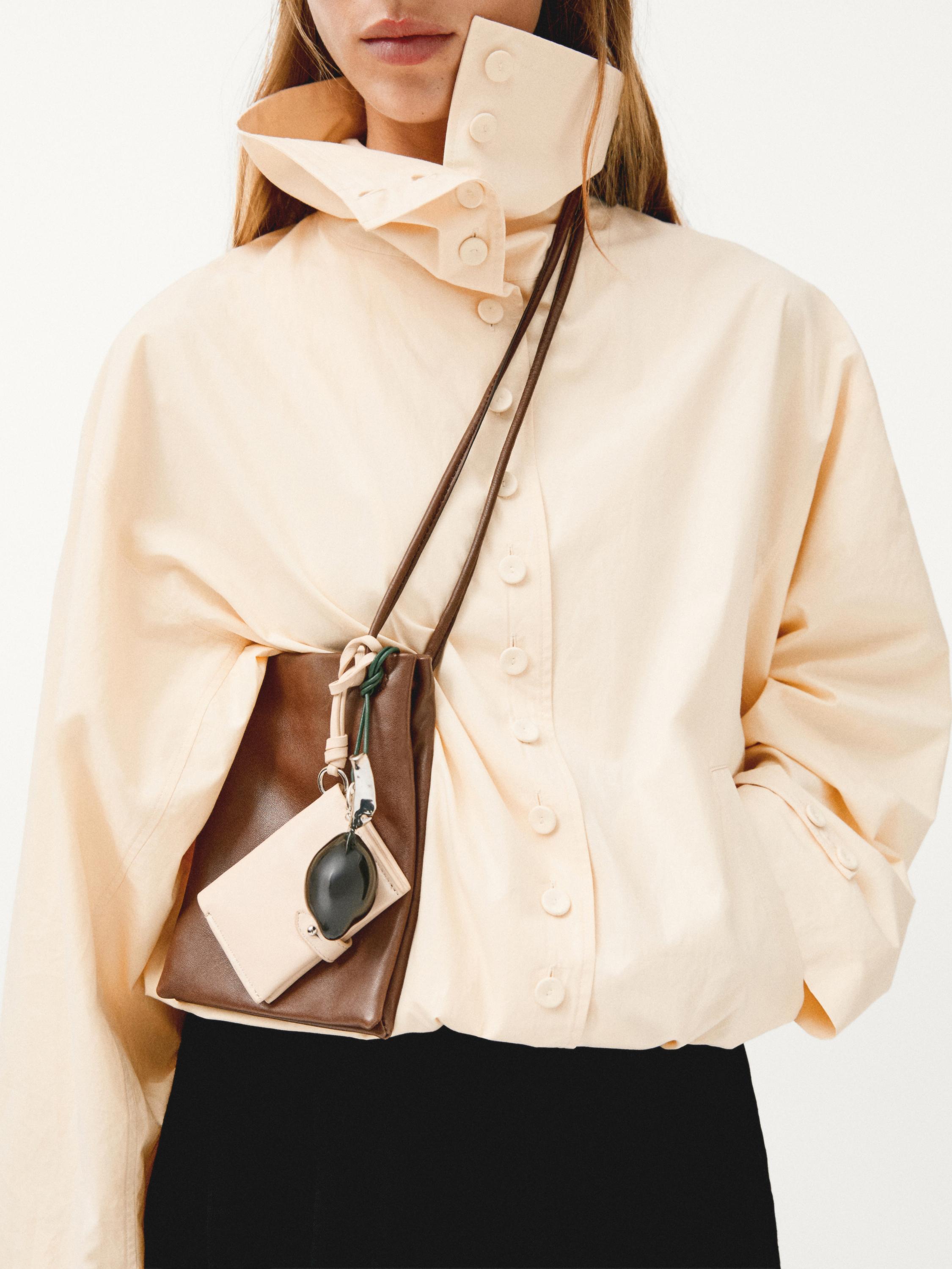 Halve close-up van een ecru jasje met opstaande kraag en knoopsluiting aan de voorkant, bruine crossbodytas met sleutelhanger en beige hoes.