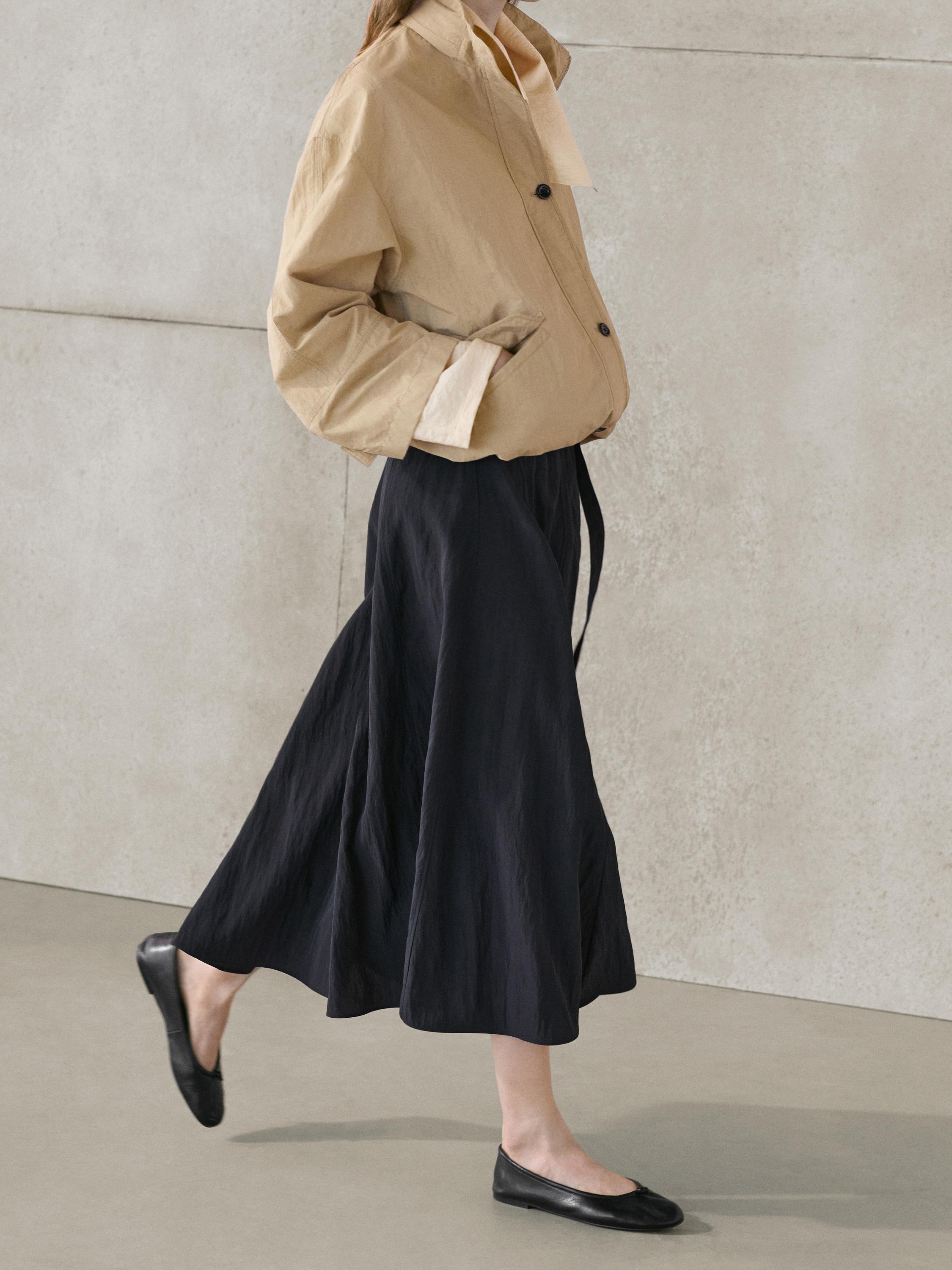 Zestaw: beżowa marynarka z guzikami i długa szaroniebieska spódnica z paskiem, czarne płaskie buty.