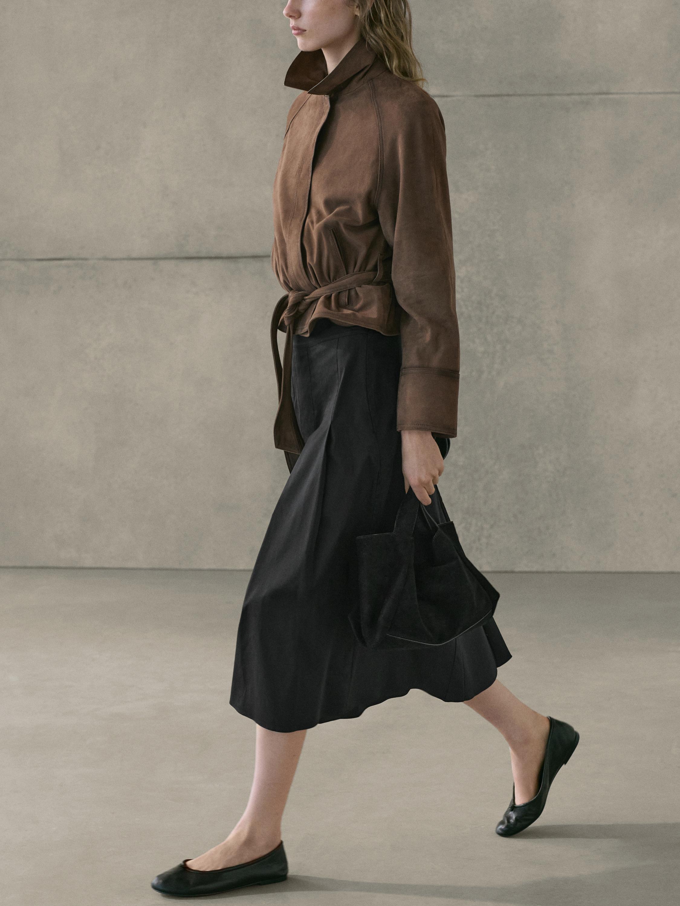 Conjunto de chaqueta marrón con cinturón y falda midi negra con tablas, bolso negro y zapatos planos negros.