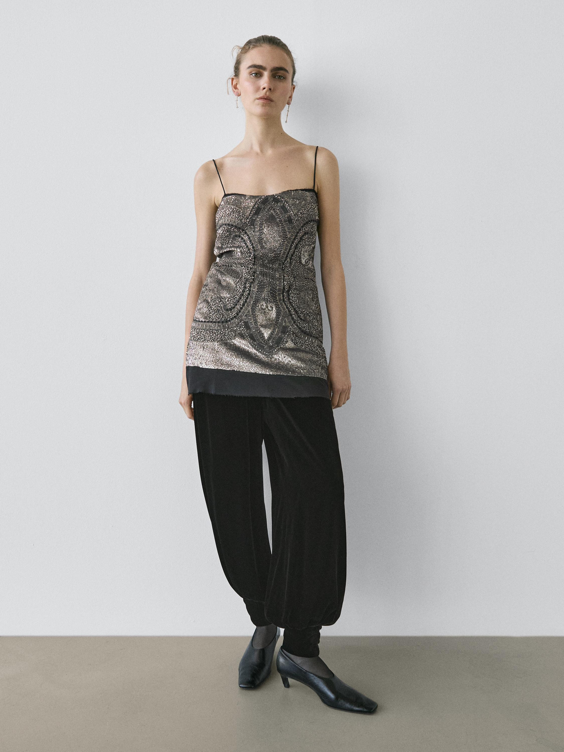 Conjunto de top plateado con tirantes delgados y bordados, pantalones negros holgados y zapatillas de tacón negras.