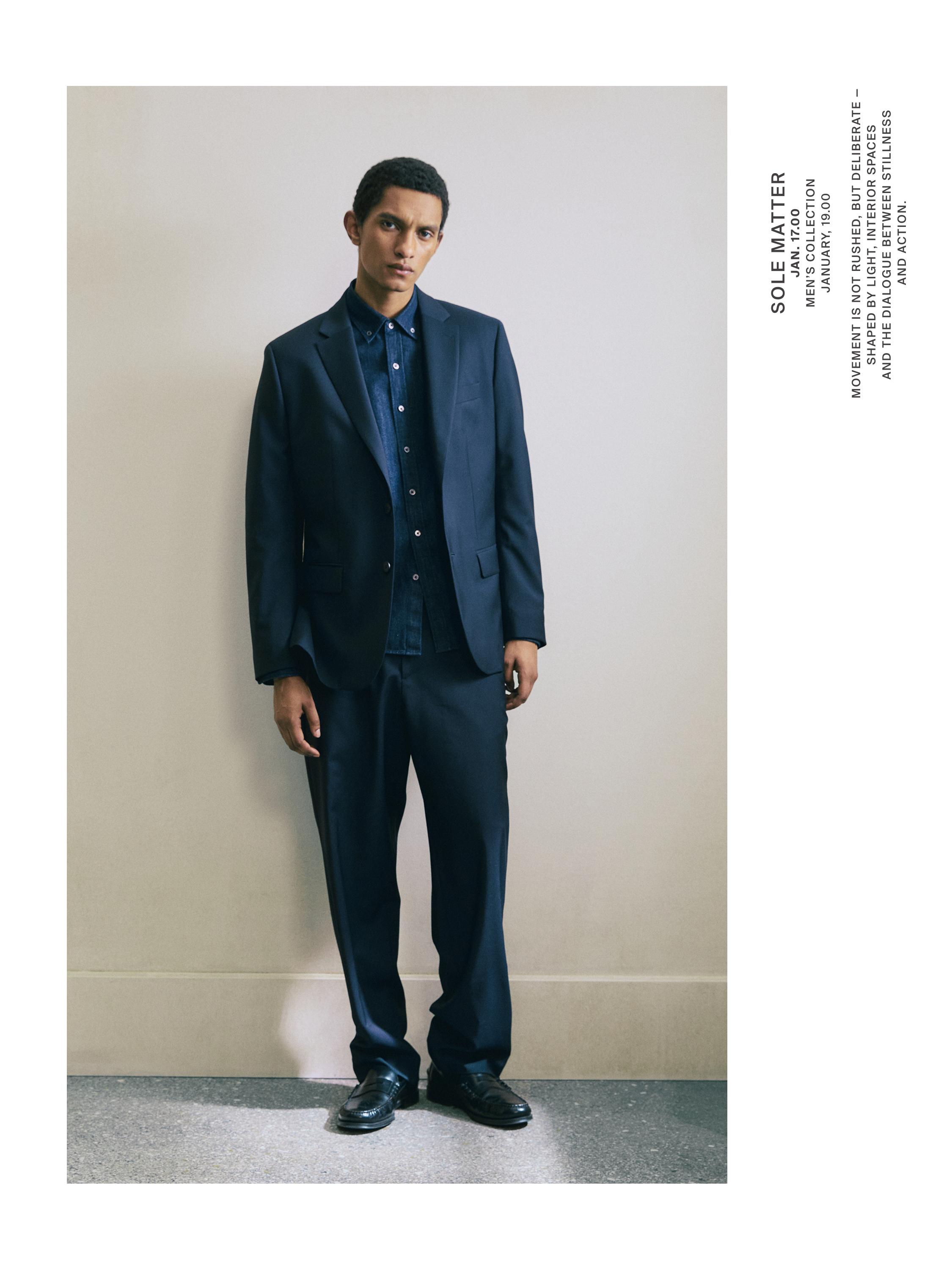 Koyu mavi takım elbise ceketi, gömleği ve pantolonu ile. Yakalı ve cepli ceket, düğmeli gömlek.