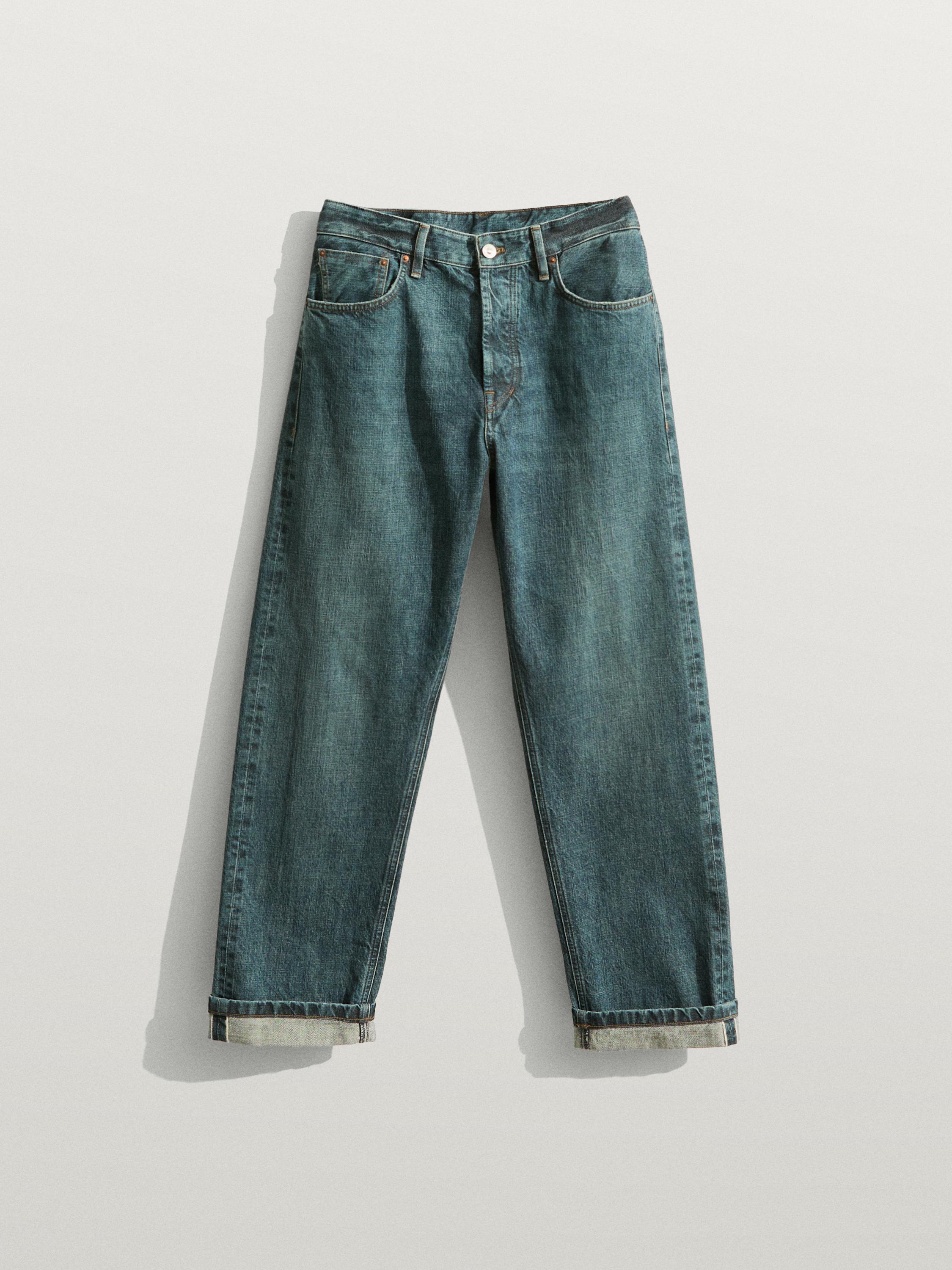 Aurrez aurreko bista: jeans indigo zuzenak, aurreko eta atzeko poltsikoekin, botoi eta kremailera itxierarekin, eta azpildura tolestuarekin.