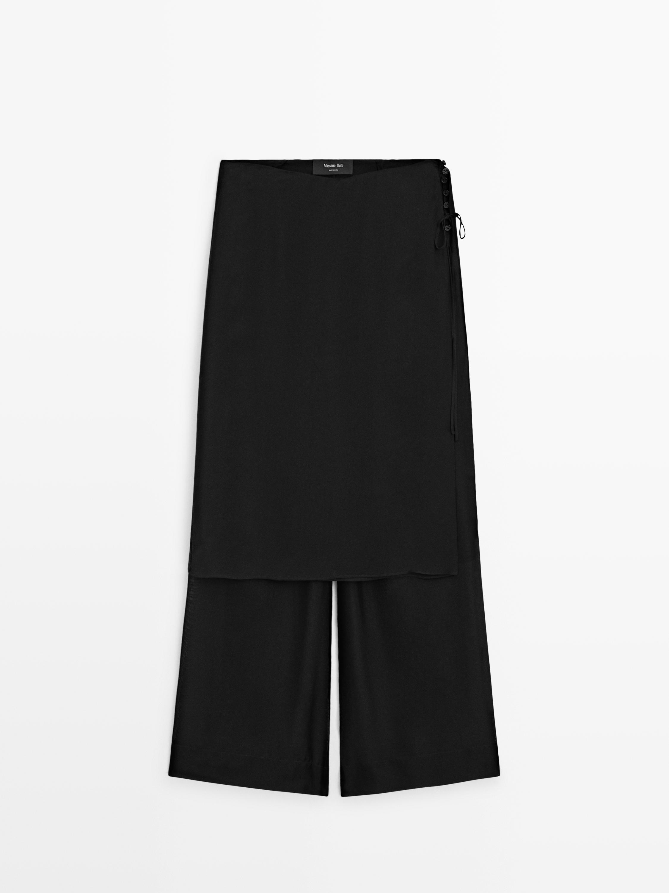 Vista frontal de calças pretas com camada sobreposta e fecho lateral de botões.