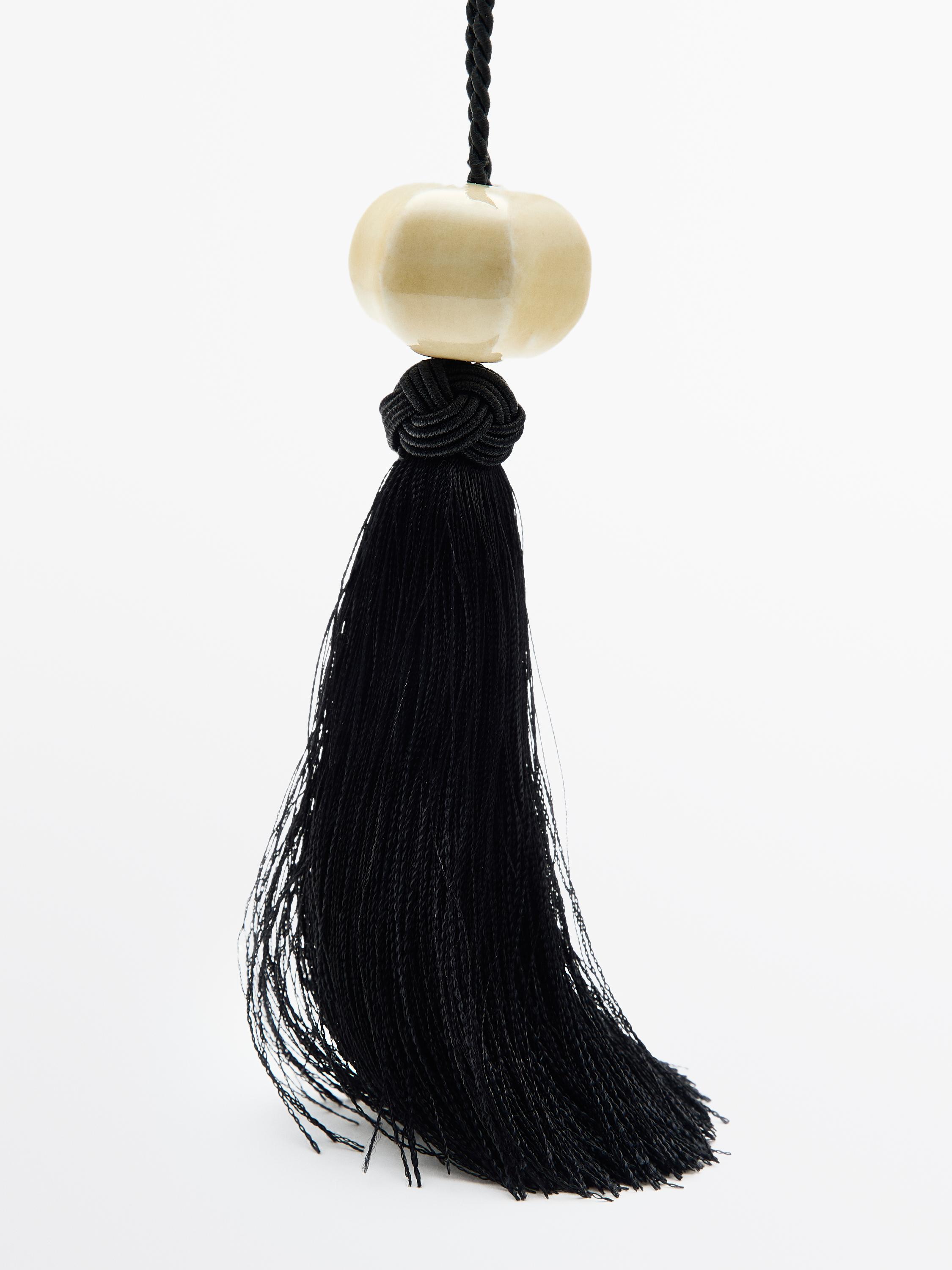Close-up of a necklace with a beige ceramic piece and a hanging black tassel, detail of braided cord at the joint.