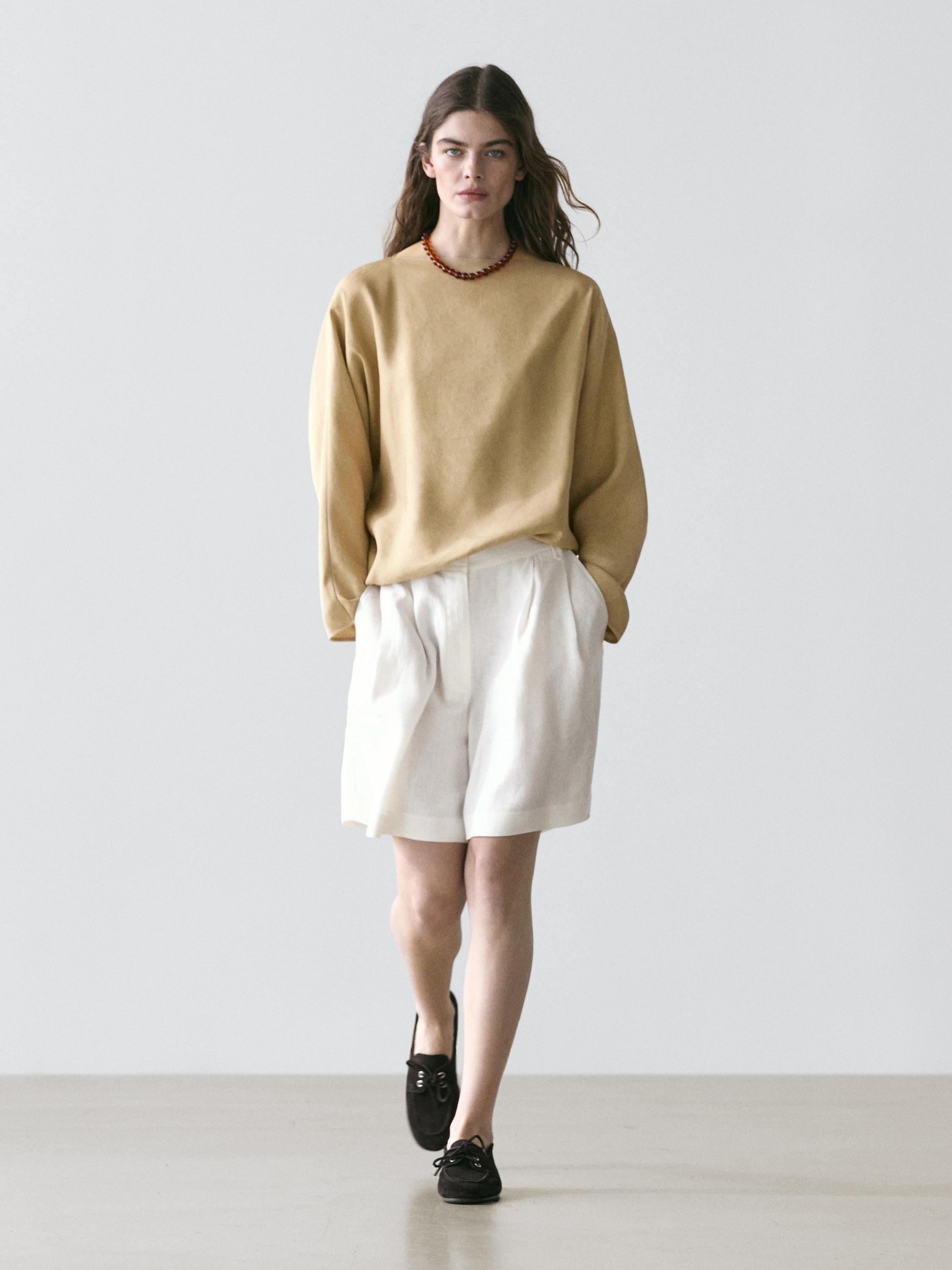 Bež džemper dugih rukava i bijele bermude s naborima, u kombinaciji s crnim cipelama.