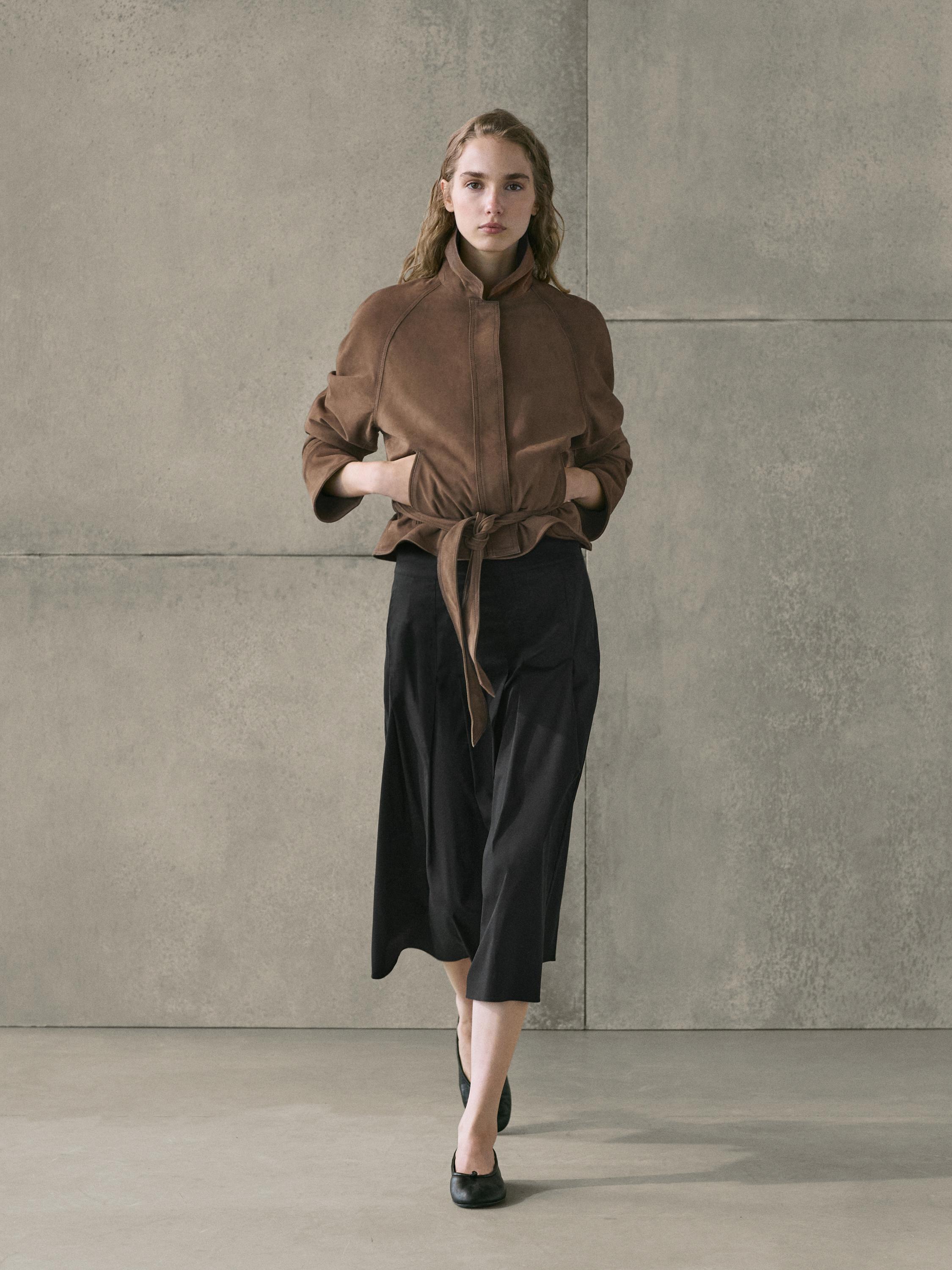 Conjunto de chaqueta marrón con cinturón y falda midi negra con tablas, combinados con zapatos negros.