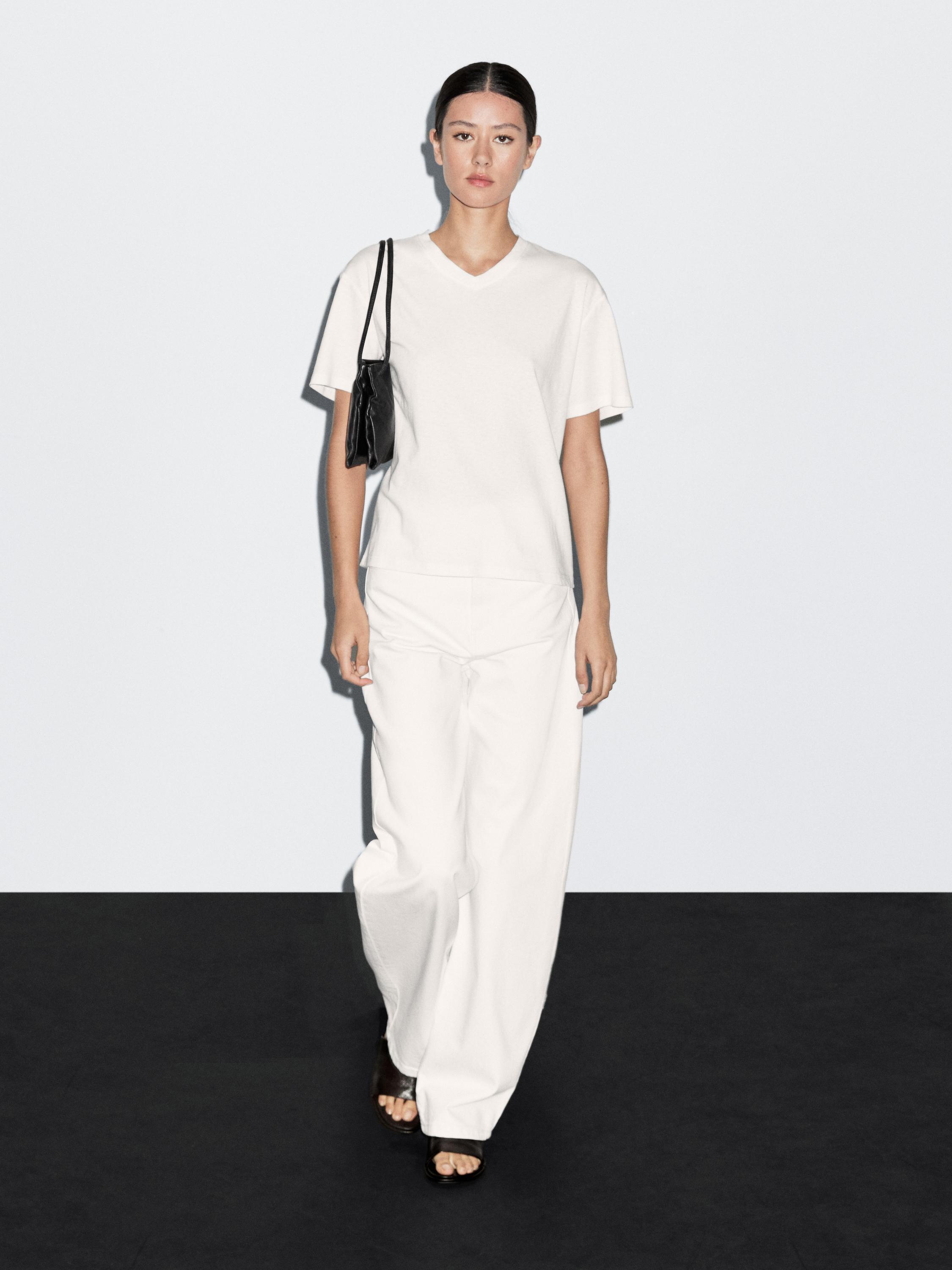 Комплект из белой футболки с коротким рукавом и белых широких брюк, черная сумка через плечо.