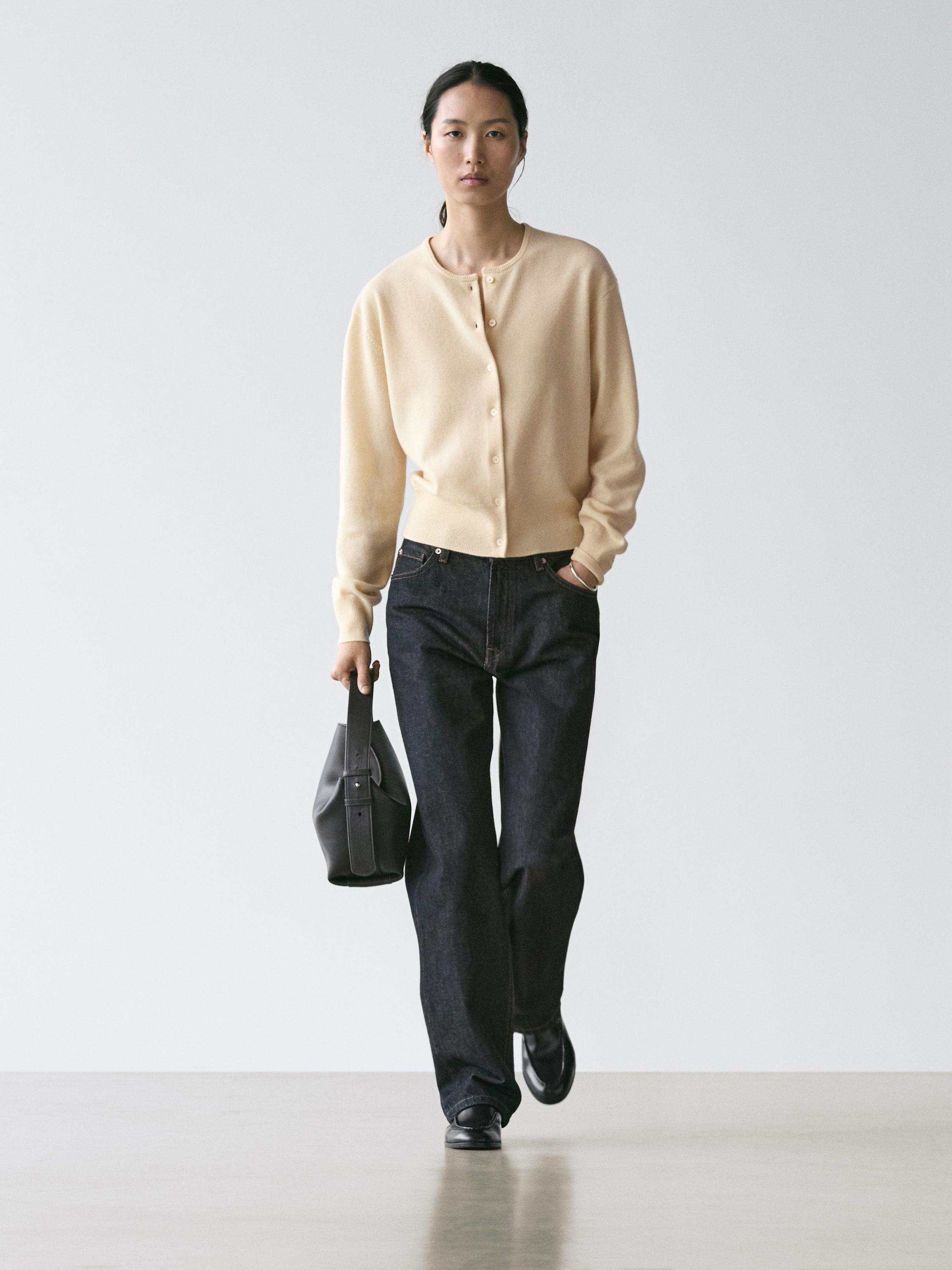 Комплект: в'язаний ванільний кардиган з круглим вирізом і ґудзиками, темні штани та чорна сумка.