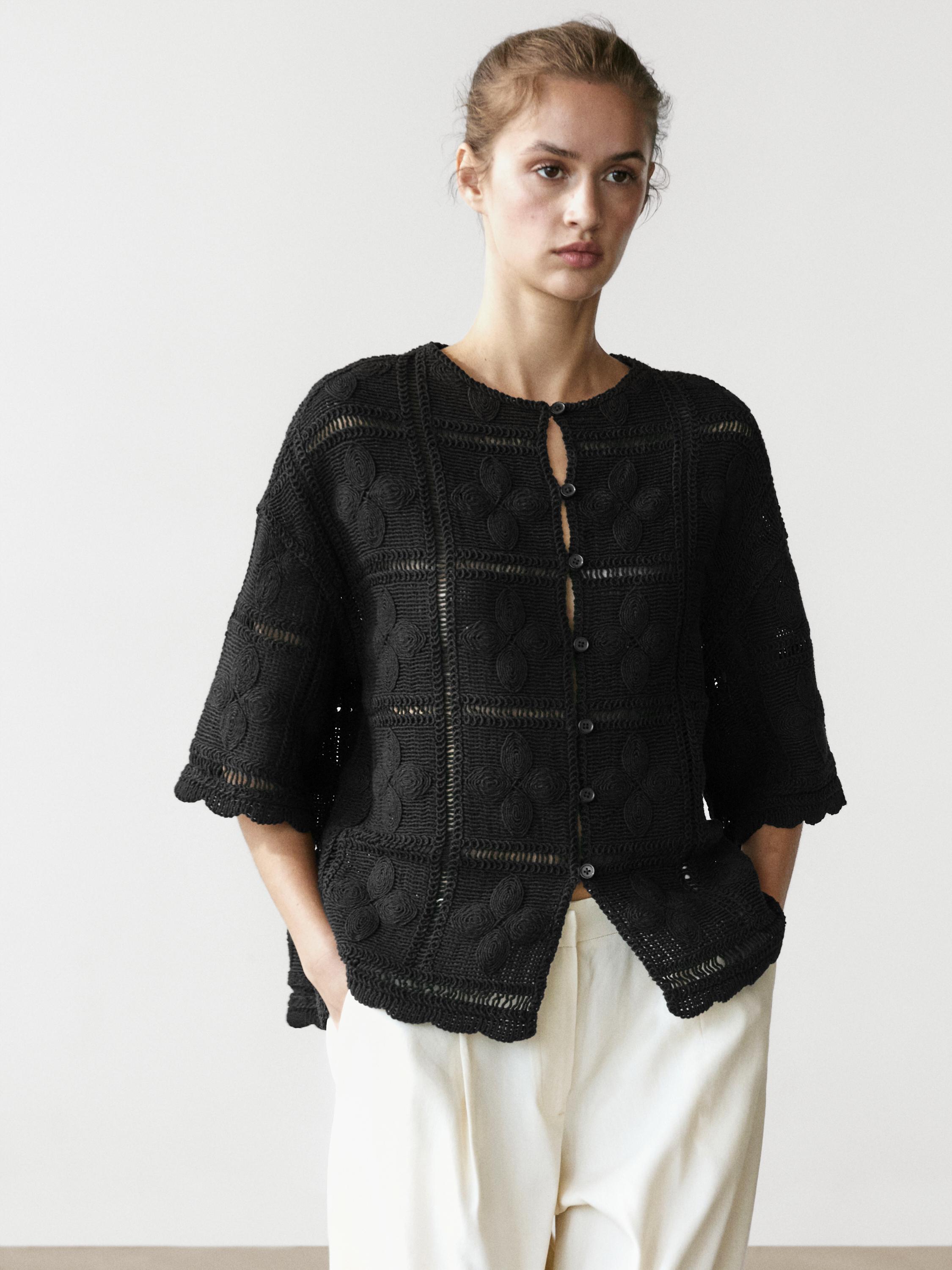 Ensemble veste noire en maille ajourée crochet avec boutons sur le devant et manches trois quarts, combinée à un pantalon clair.