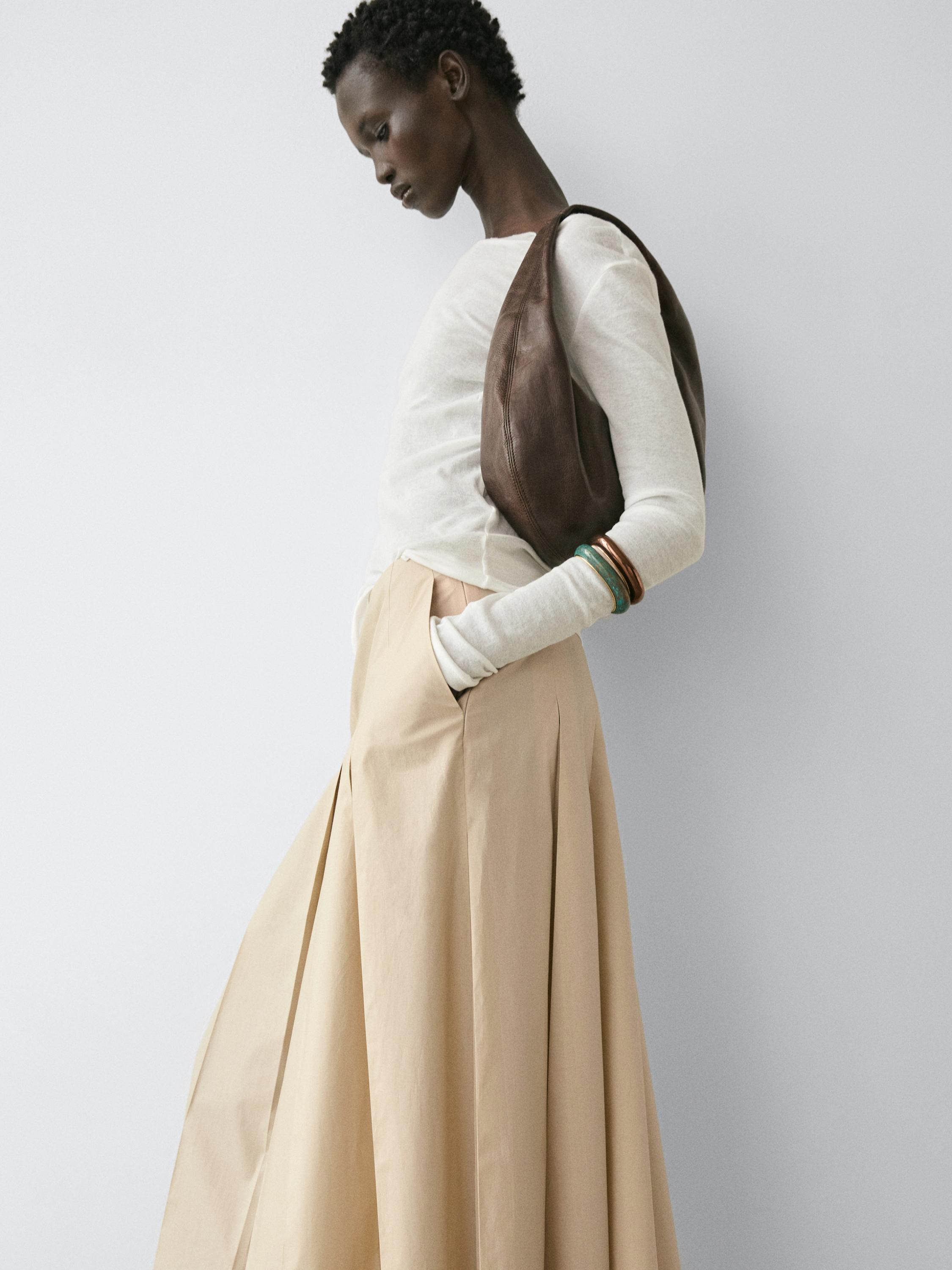 Plano medio de falda midi arena con tablas y bolsillos laterales, combinada con blusa blanca de manga larga.