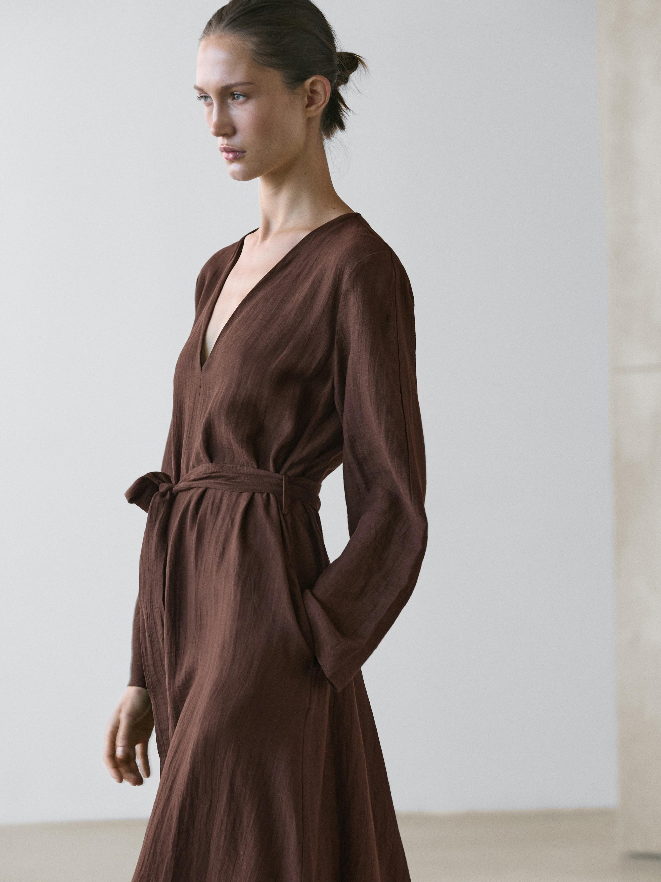 Μεσαίο πλάνο ενός ρευστού μίντι φορέματος σε καφέ χρώμα με ζώνη, V-λαιμόκοψη και μακριά μανίκια, με πλαϊνές τσέπες.