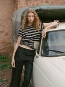 Model met een gestreept T-shirt en een donkere broek, leunend tegen een witte auto met een achtergrond van een aarden muur.