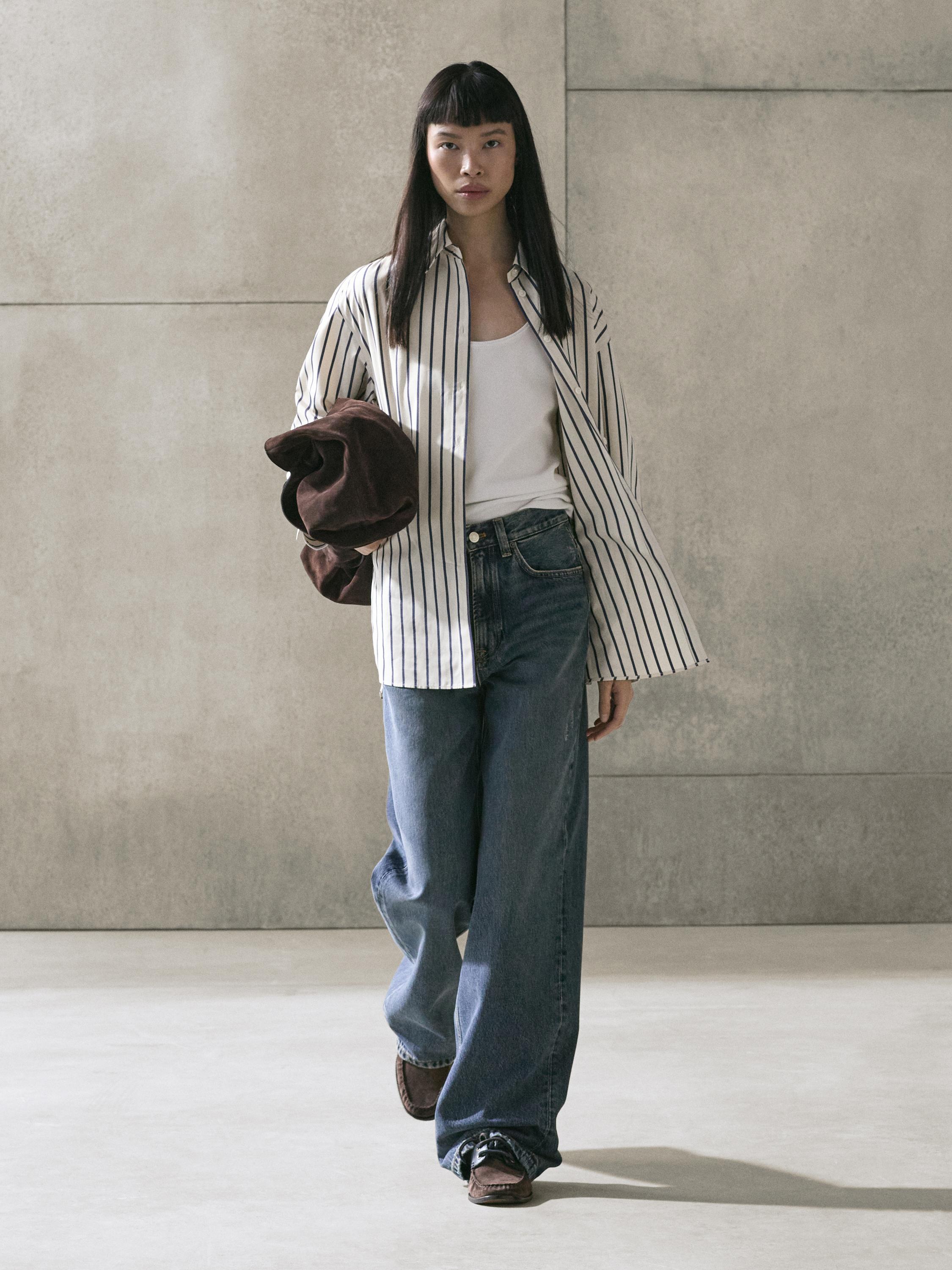 Completo di camicia a righe color vaniglia, maglietta bianca, jeans larghi blu e mocassini marroni.