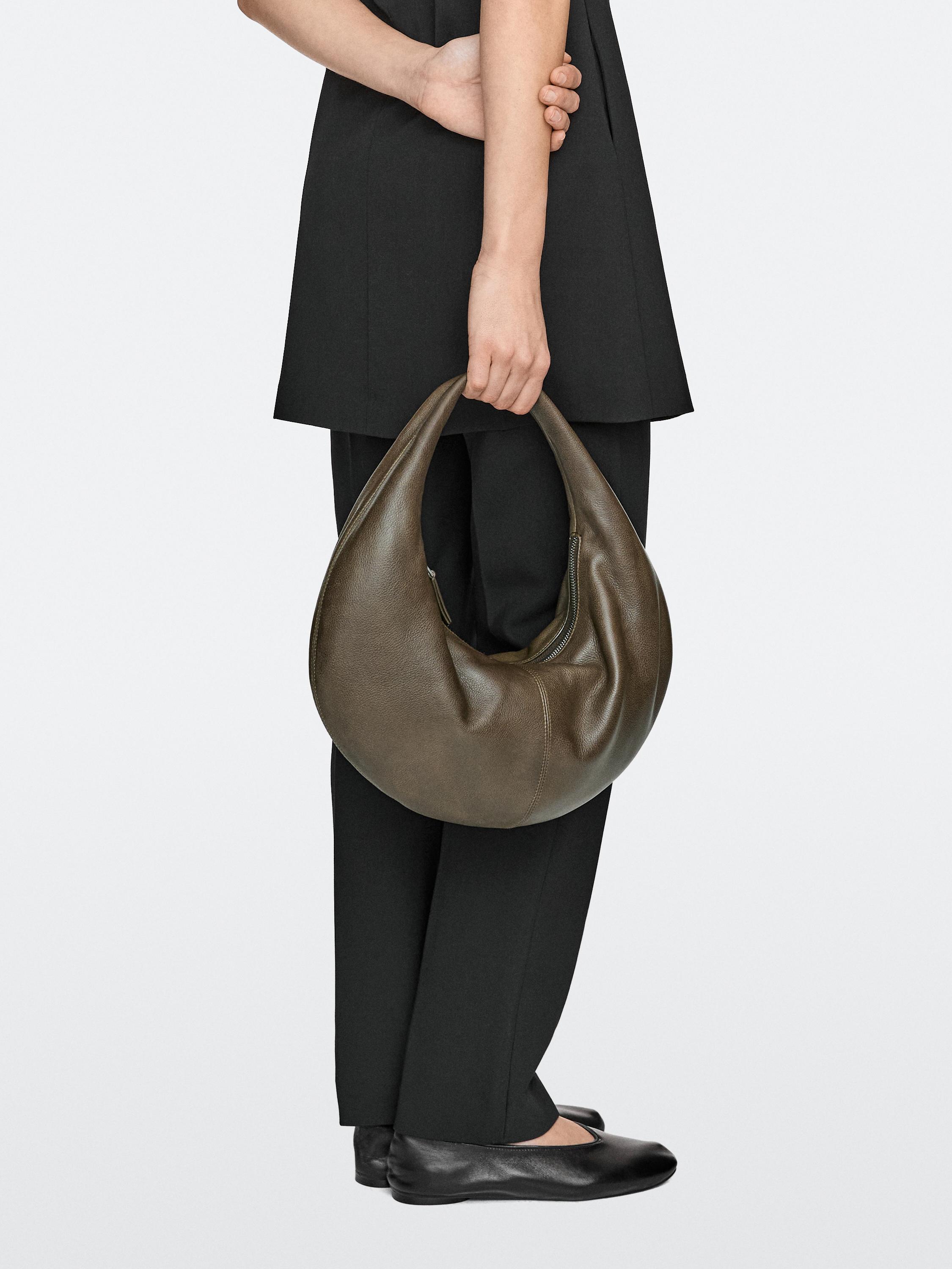 Set aus olivgrüner Handtasche mit geschwungenem Design, in der Hand getragen, kombiniert mit schwarzer Hose und schwarzen Schuhen.