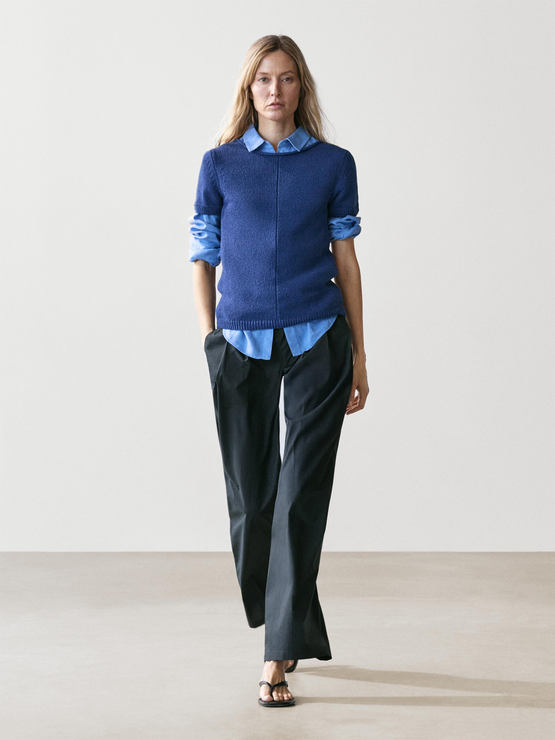 Beli bol paça lacivert pantolon ve açık mavi gömleğin üzerine katlanmış kollu koyu mavi kazak.