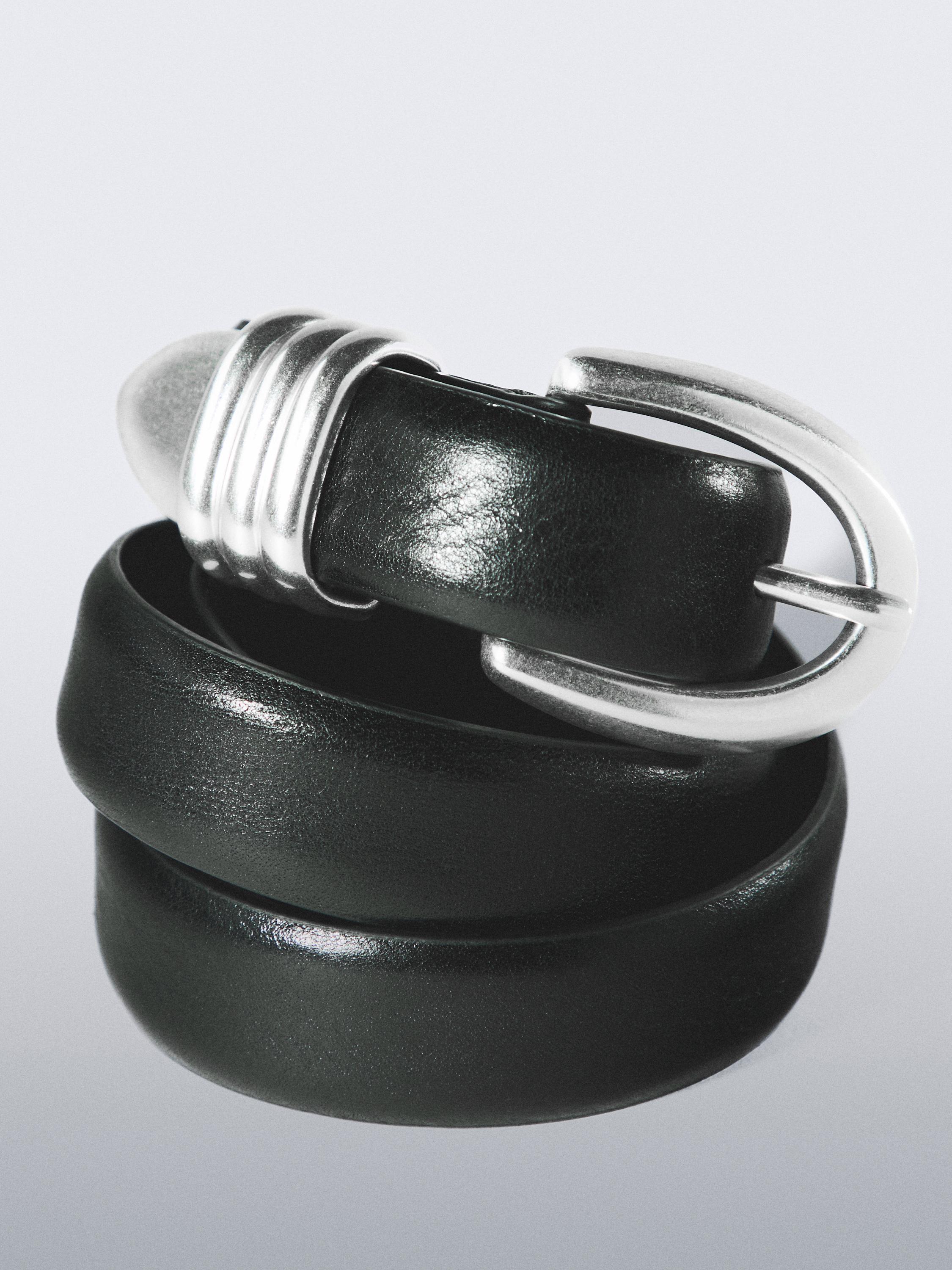 Cinturón negro con hebilla metálica plateada y detalle decorativo en el extremo.