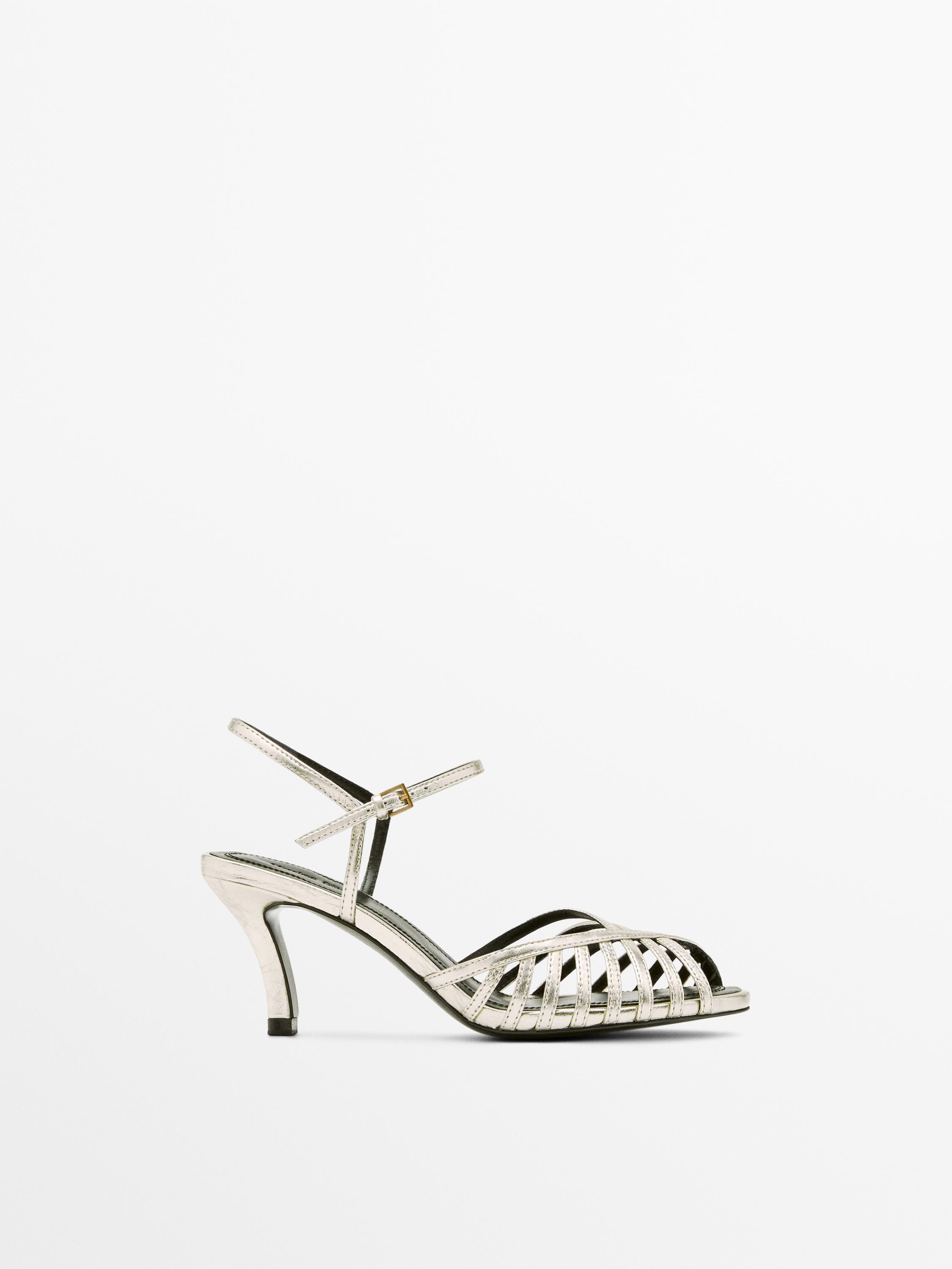 منظر جانبي لصندل بكعب عالٍ بلون البلاتيني مع أشرطة متقاطعة وإبزيم إغلاق عند الكاحل.