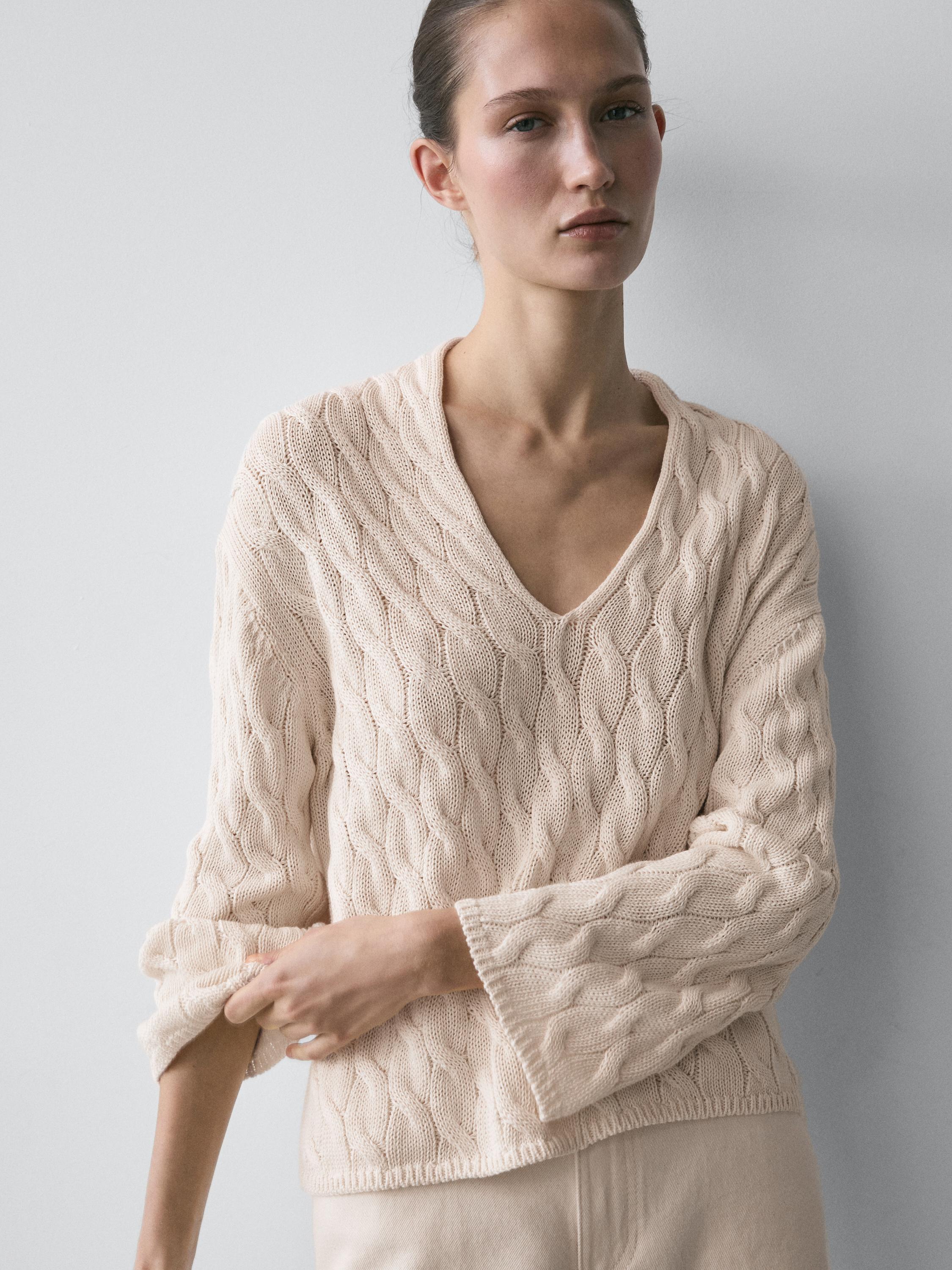Plano medio di maglione ecru con scollo a V e dettaglio a nodi. Maniche lunghe e texture intrecciata visibile.