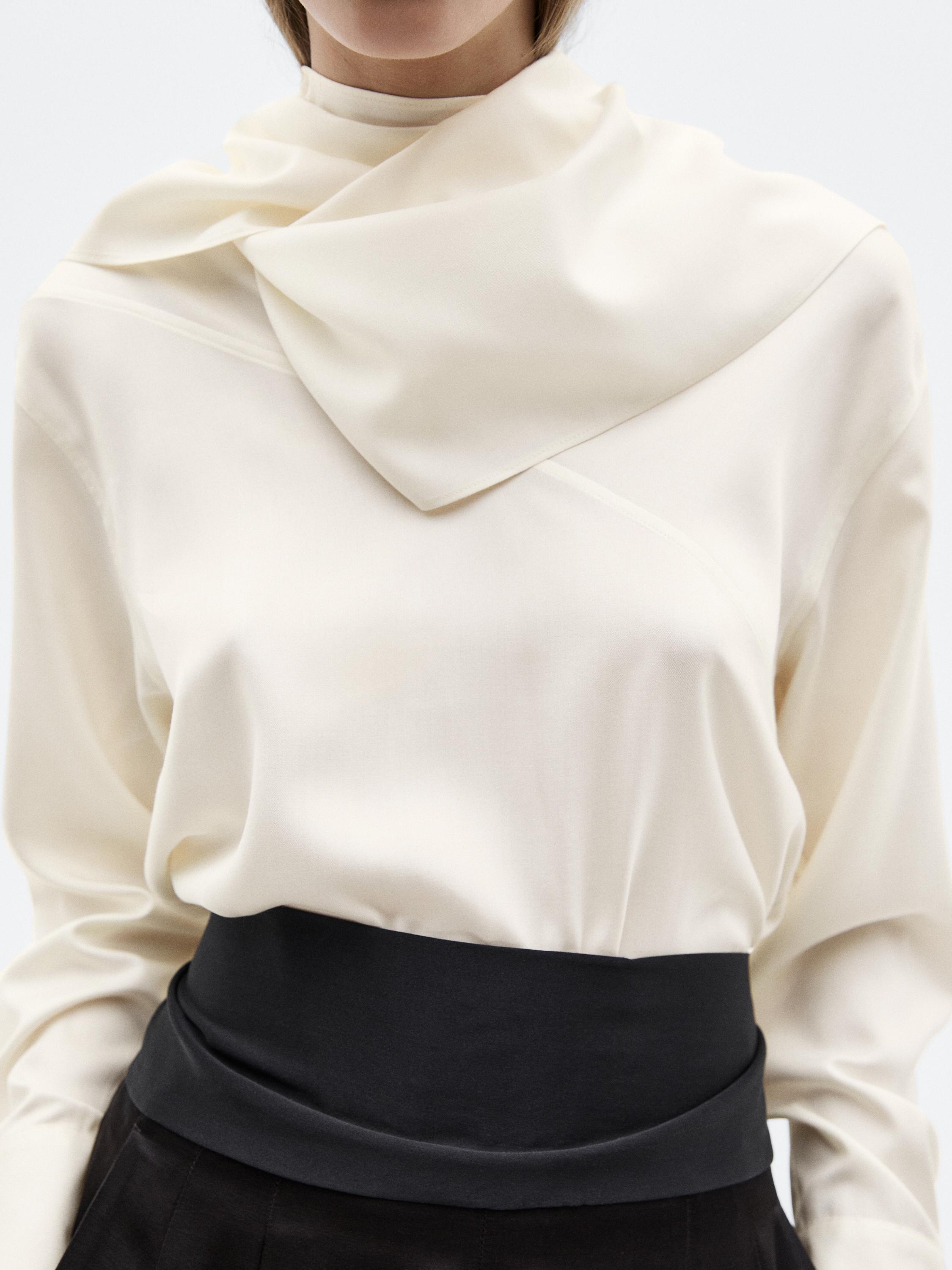 Halbaufnahme einer weißen Bluse mit drapiertem Kragen und breitem schwarzem Gürtel.