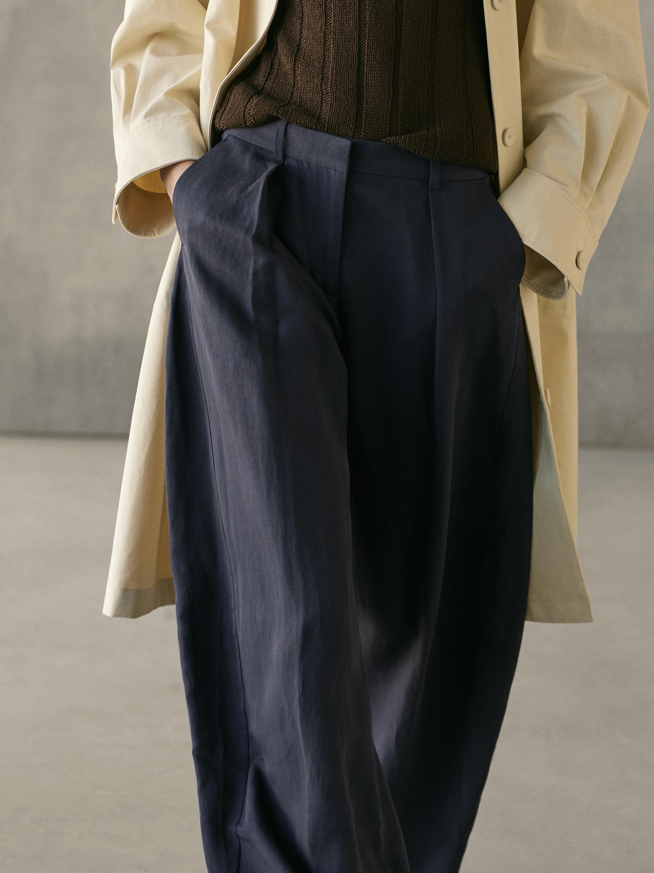 Keskmine plaan voolavatest tumesinistest pükstest, millel on voldid ja küljetaskud, kombineeritud beeži mantli ja pruuni kampsuniga.