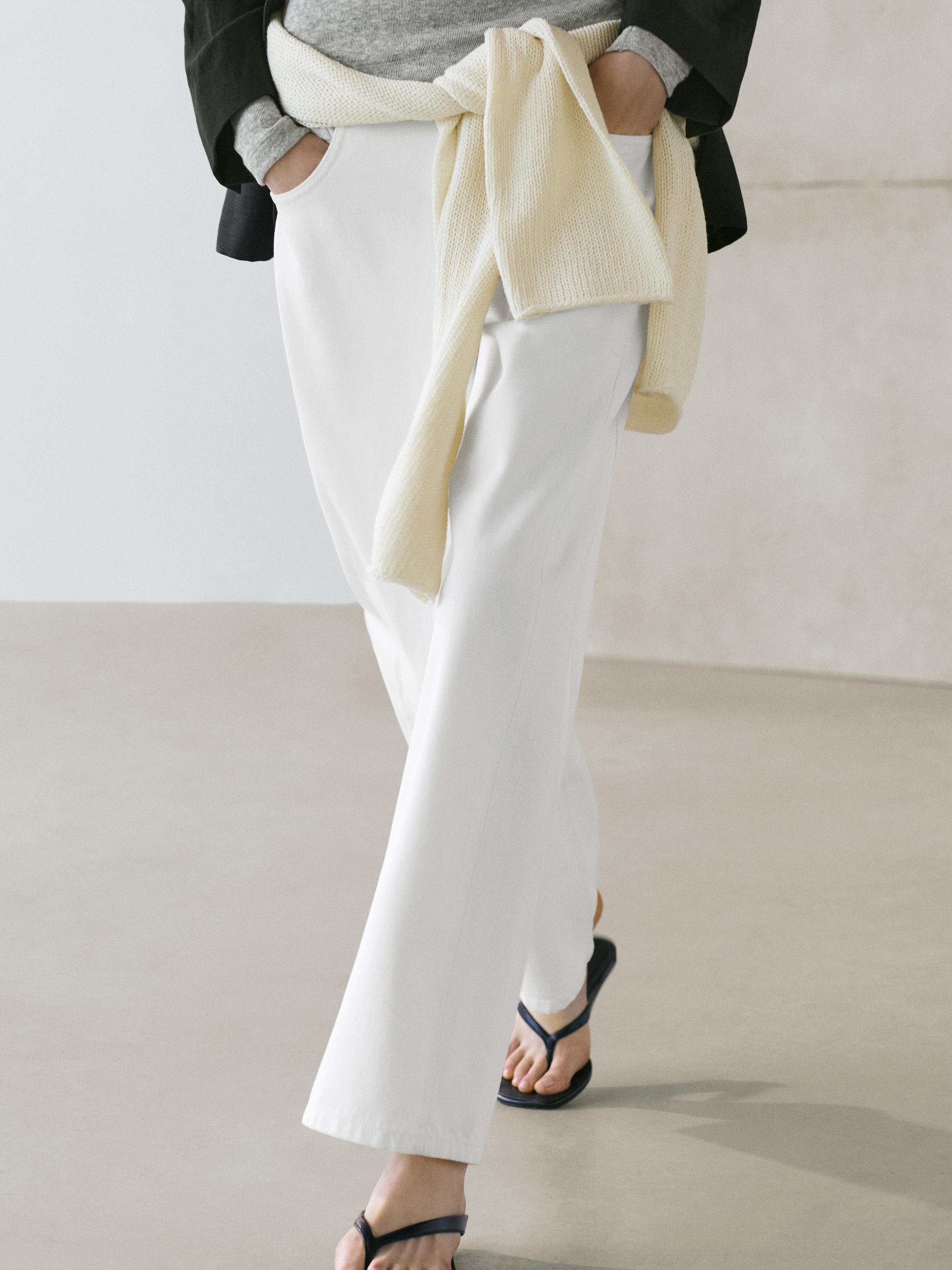 Mid-shot of white pants with side pockets, a beige sweater tied at the waist, and black sandals.