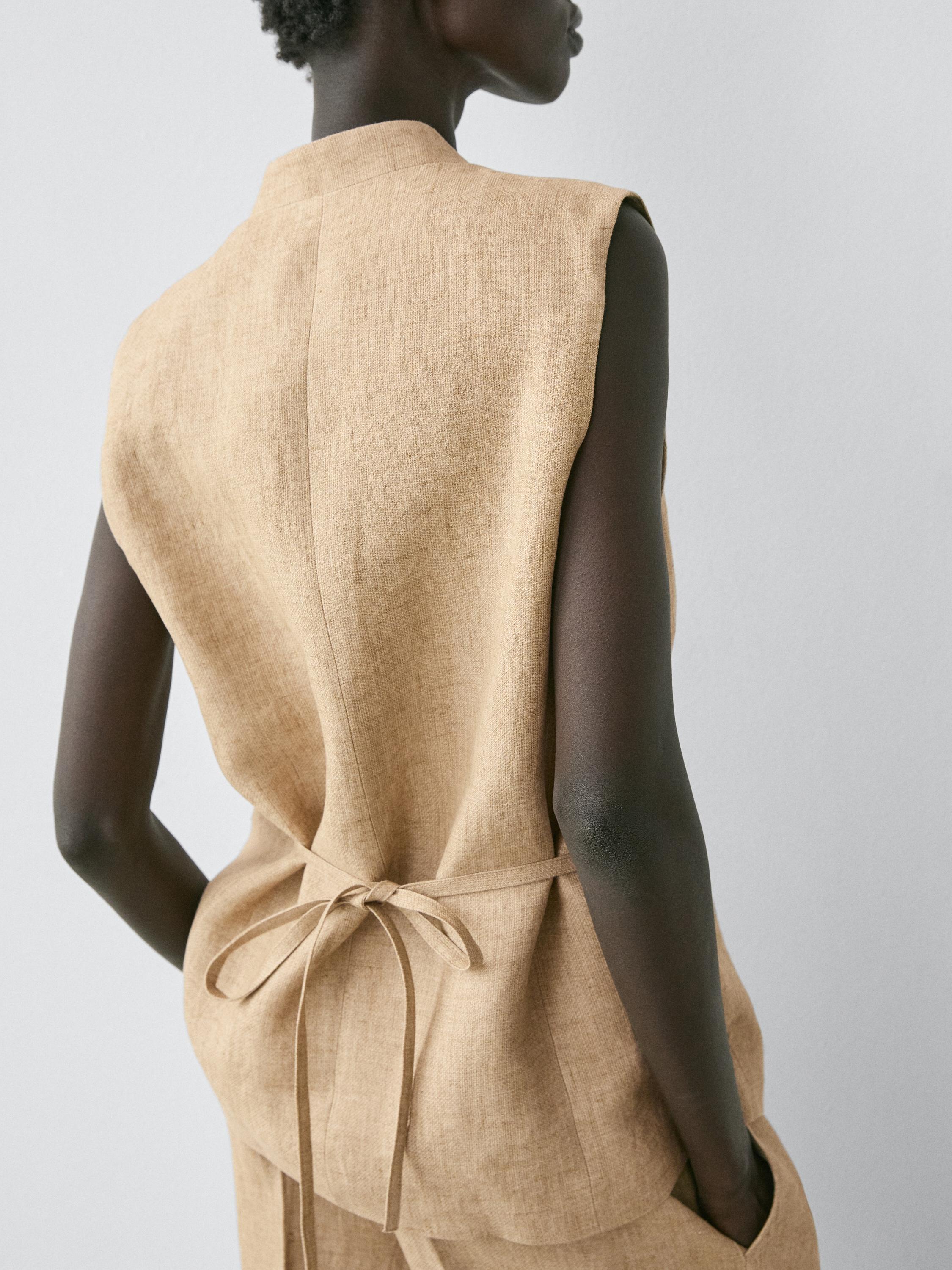 Plano medio da parte traseira dun chaleco beis sen mangas con peche cruzado e lazo na cintura.